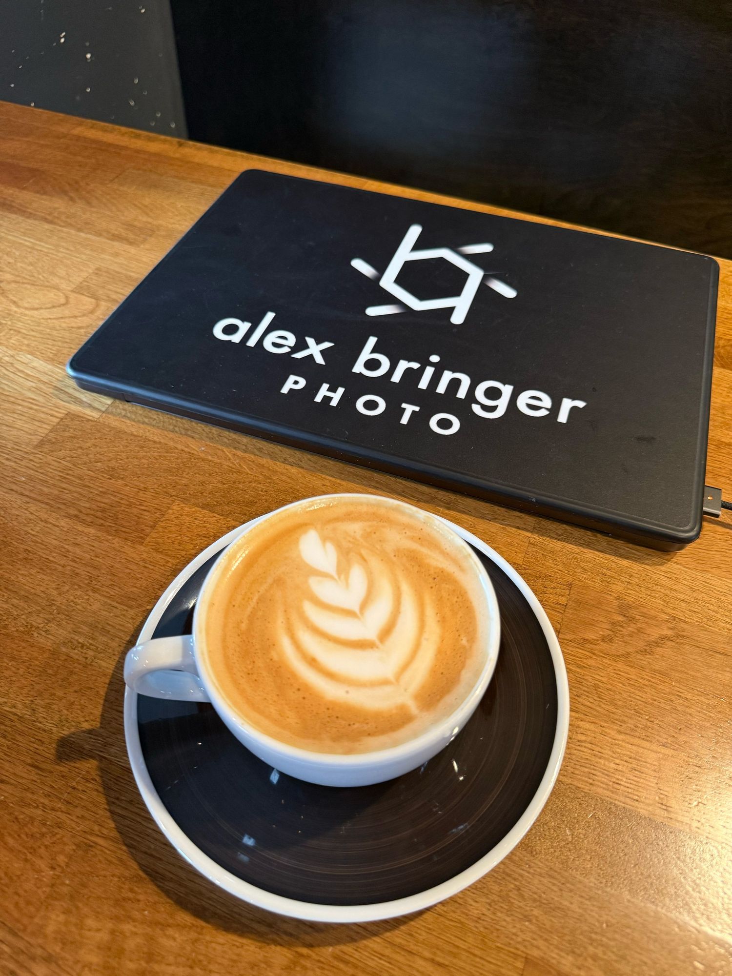Artisan latte with leaf art in white cup beside an Alex Bringer Photo branded business card on wood table.