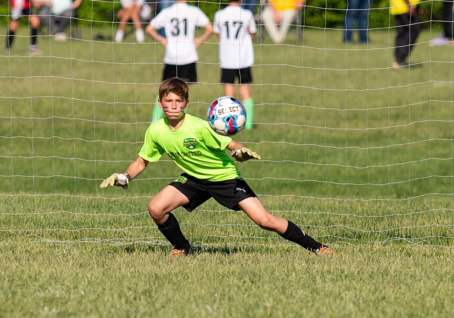 Young soccer goalkeeper in neon green jersey diving to save a ball during a youth match on a grass field.