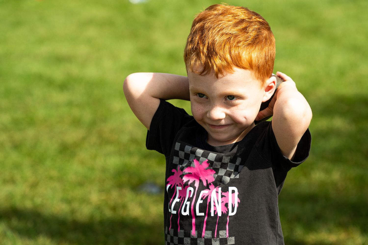 Cute red-haired boy in black t-shirt smiling on green grass field.