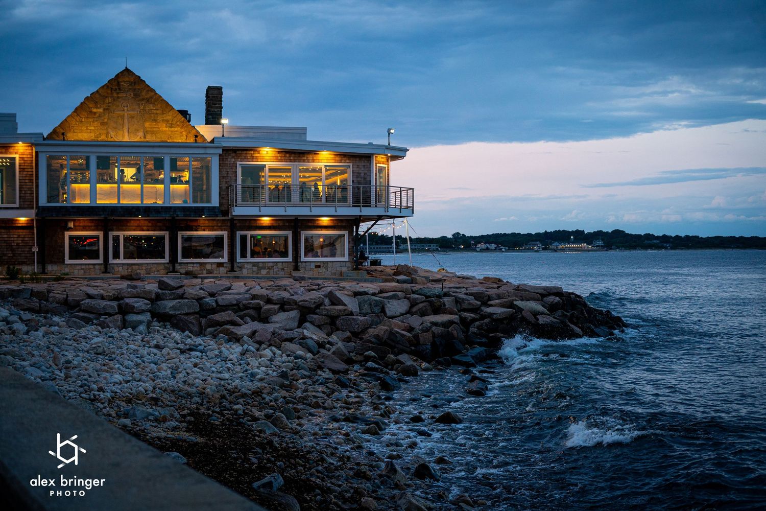 Waterfront restaurant with warm lights reflecting on rocky shoreline at dusk.