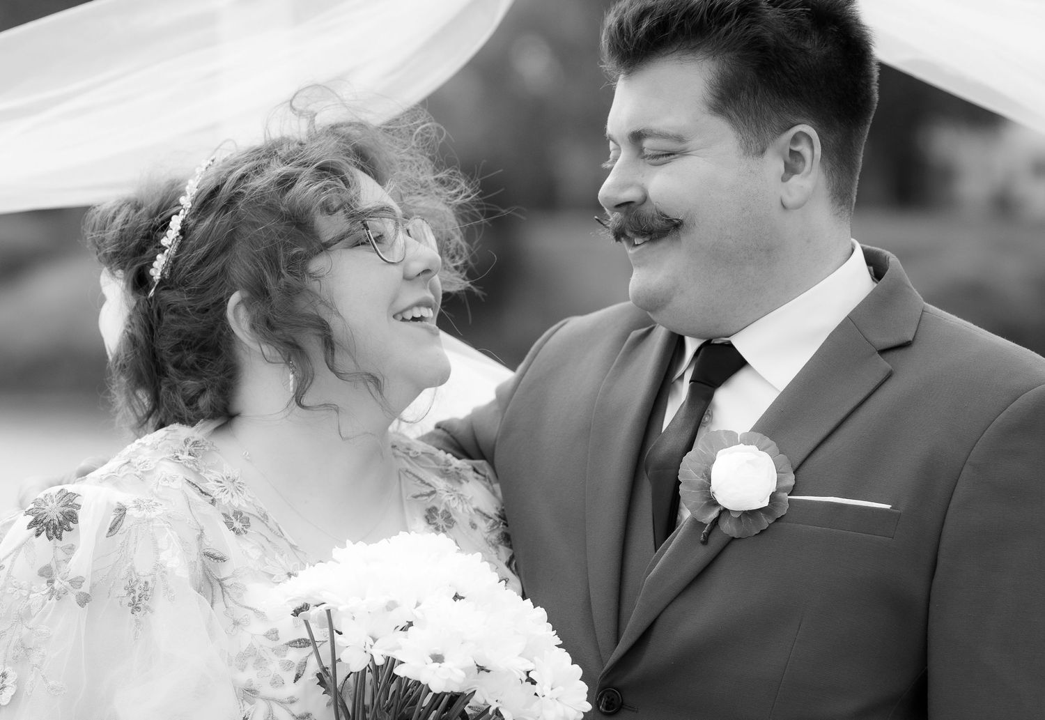 Black and white wedding photo of smiling bride with bouquet and groom with mustache in suit sharing a joyful moment.