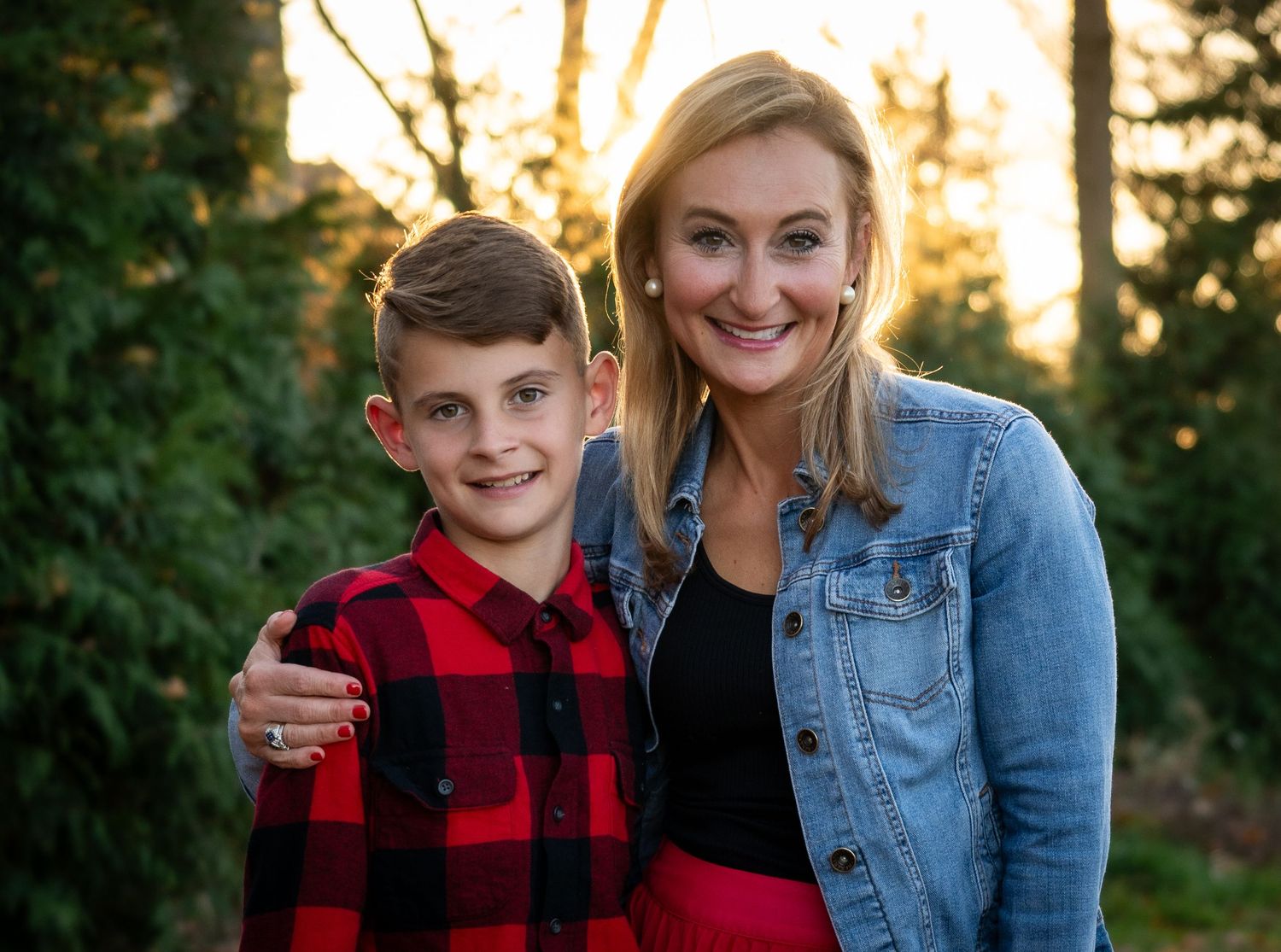 Smiling boy in red plaid shirt poses with blonde woman in denim jacket during fall outdoor portrait.