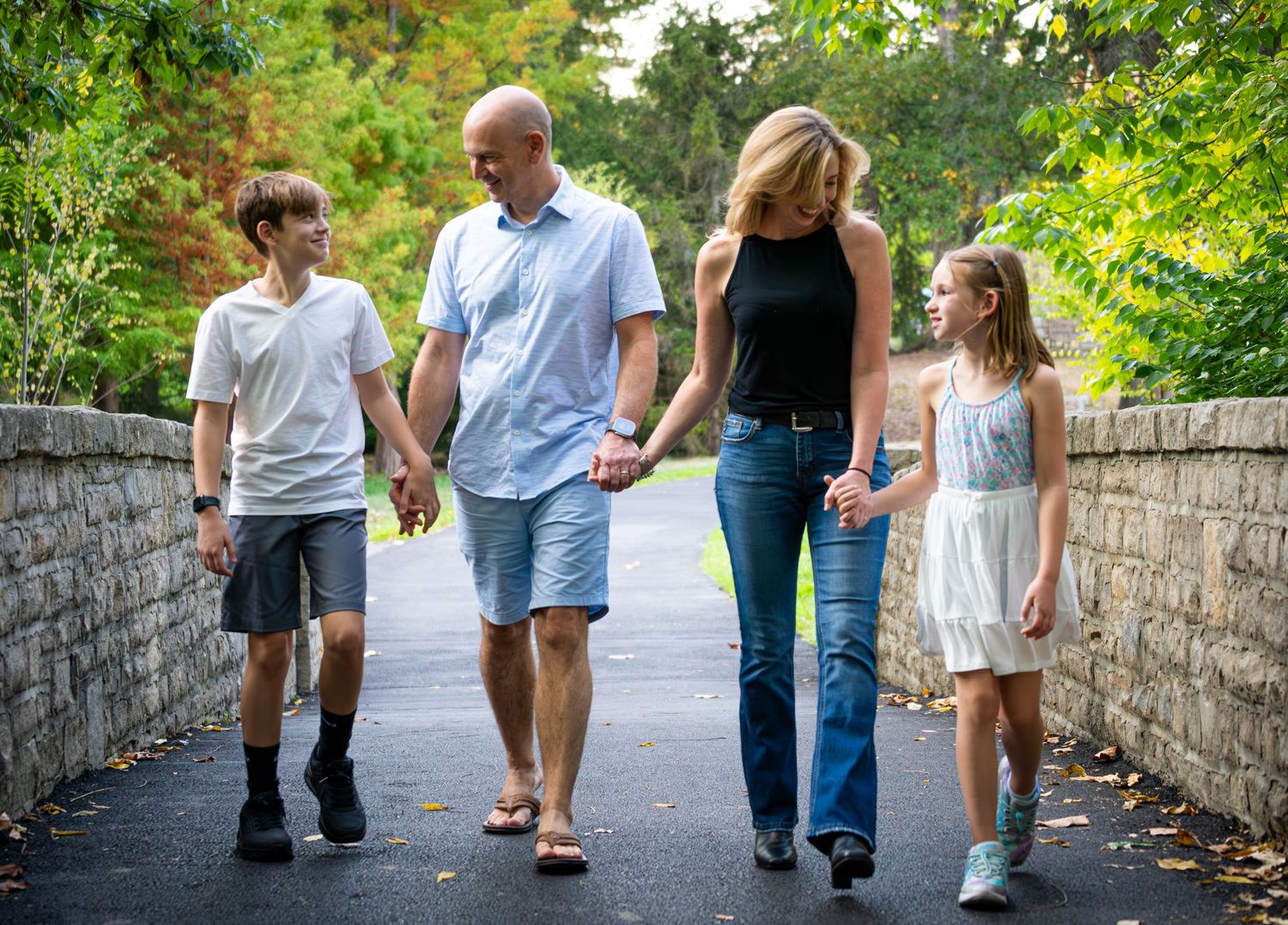 Family of four walking hand-in-hand along a scenic tree-lined pathway during a casual outdoor stroll.