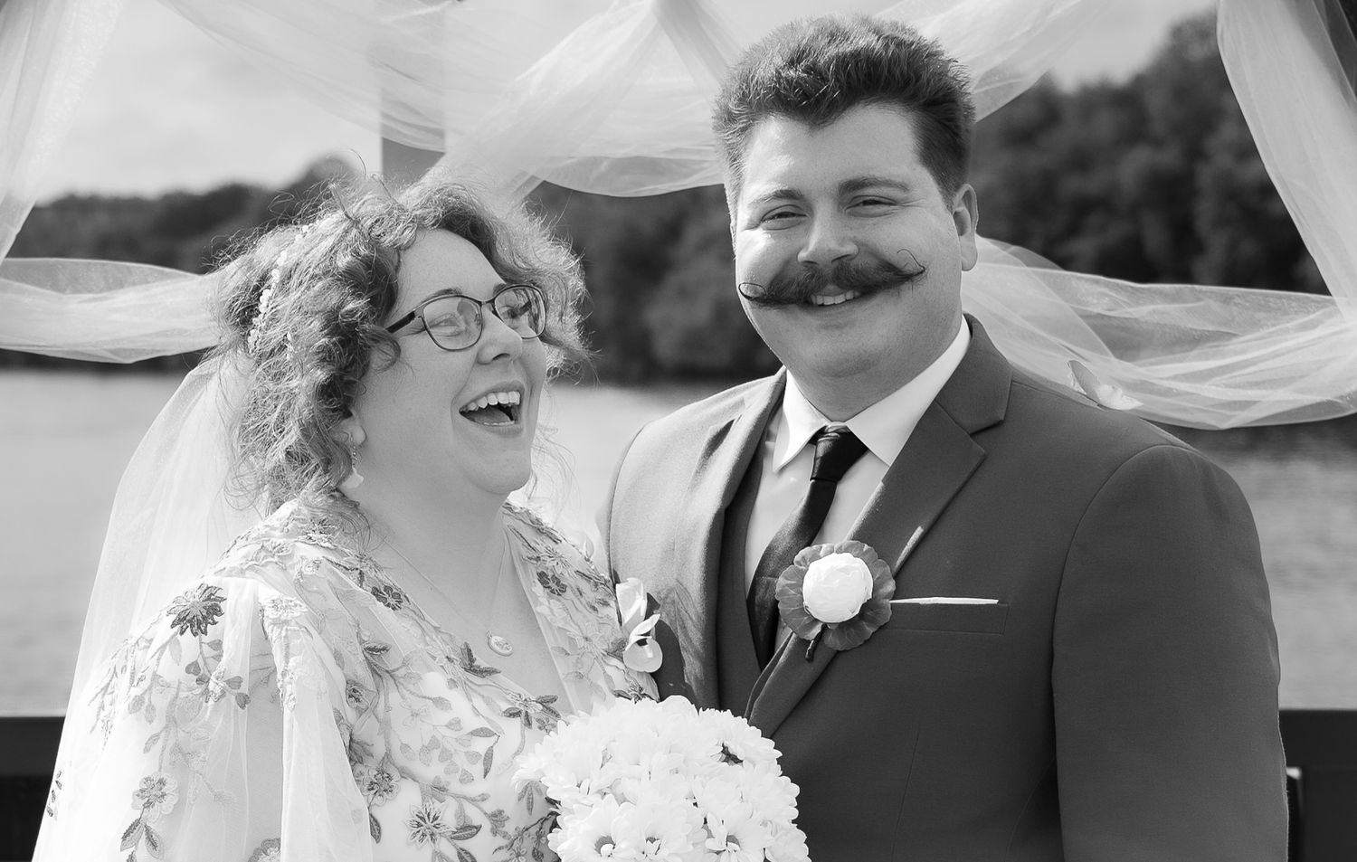 Black and white wedding photo of smiling bride with glasses holding bouquet and groom with mustache in suit.