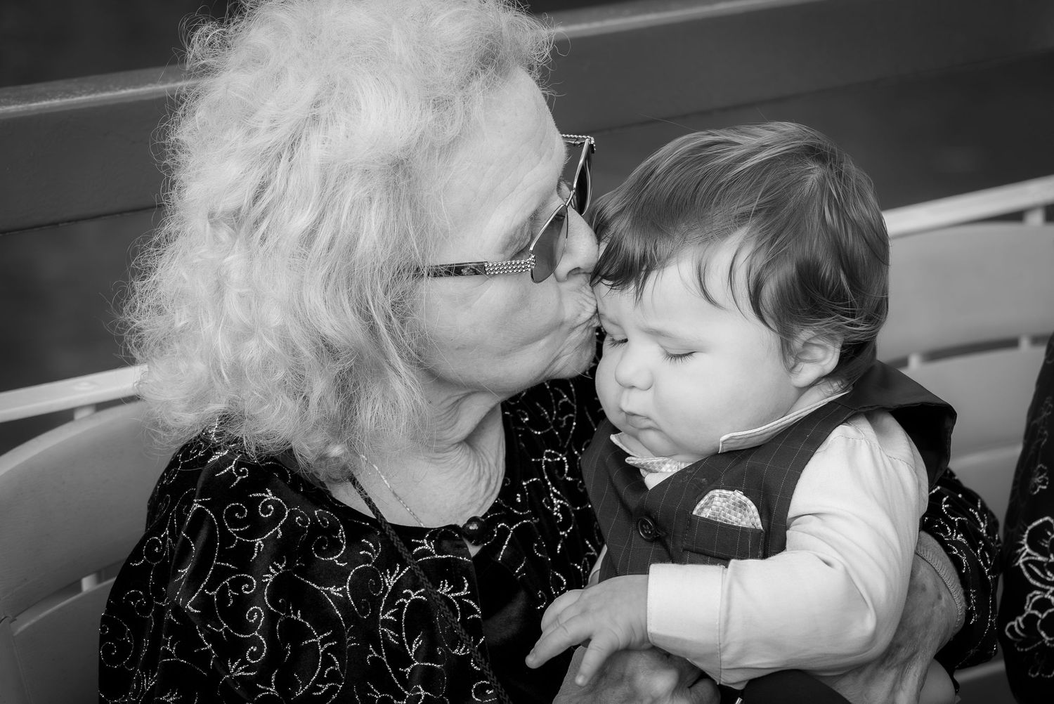 Elderly woman with curly white hair and glasses kissing a cute baby in a black and white portrait photo.
