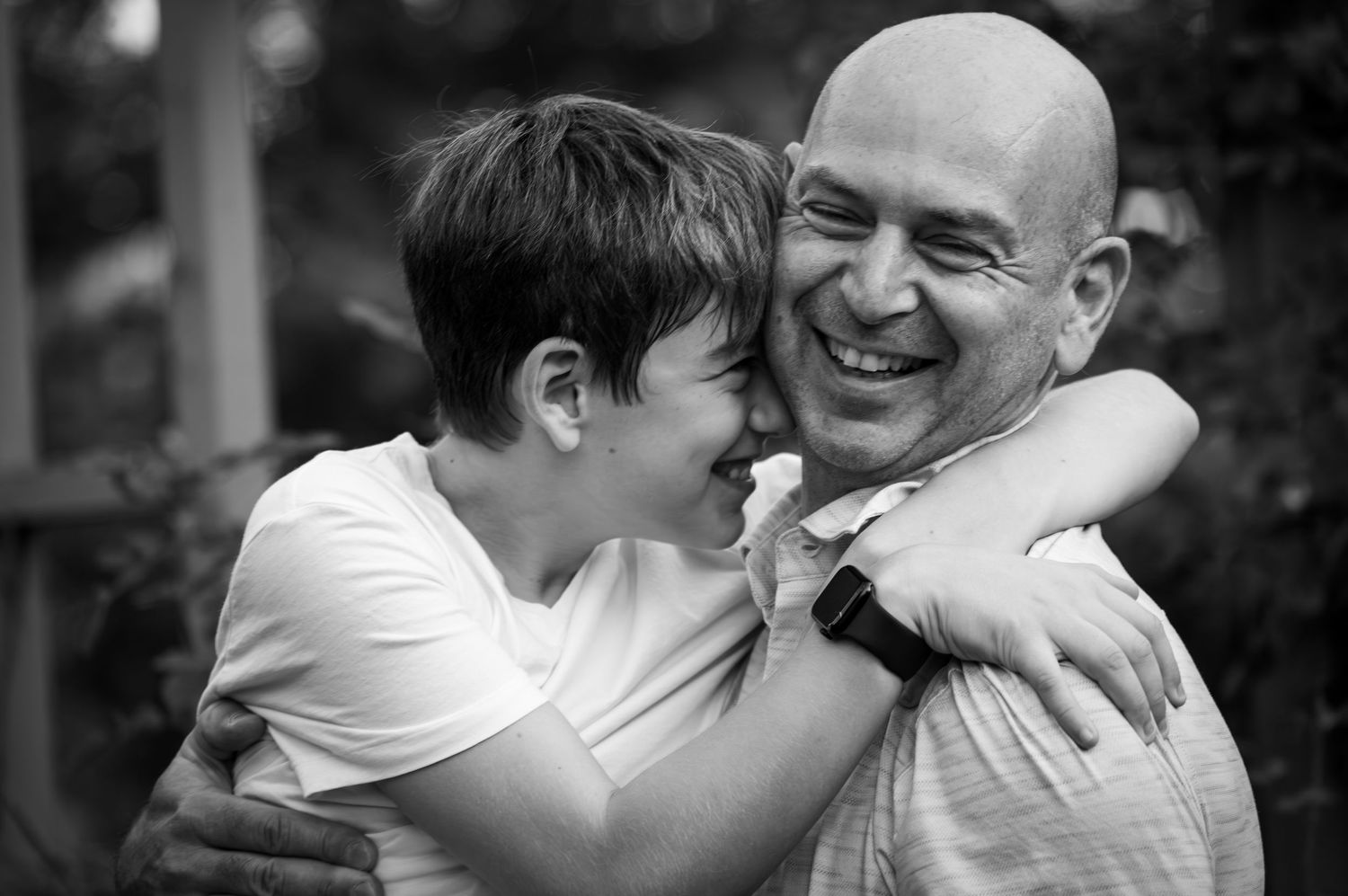 Black and white photo of bald man laughing warmly while being hugged by young boy outdoors.