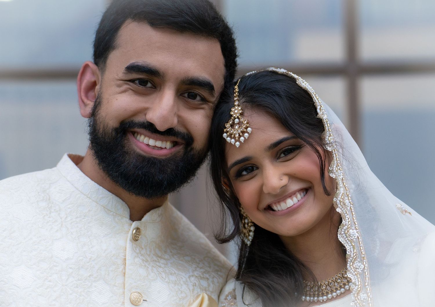 South Asian couple in traditional white wedding attire smiling together on their wedding day.