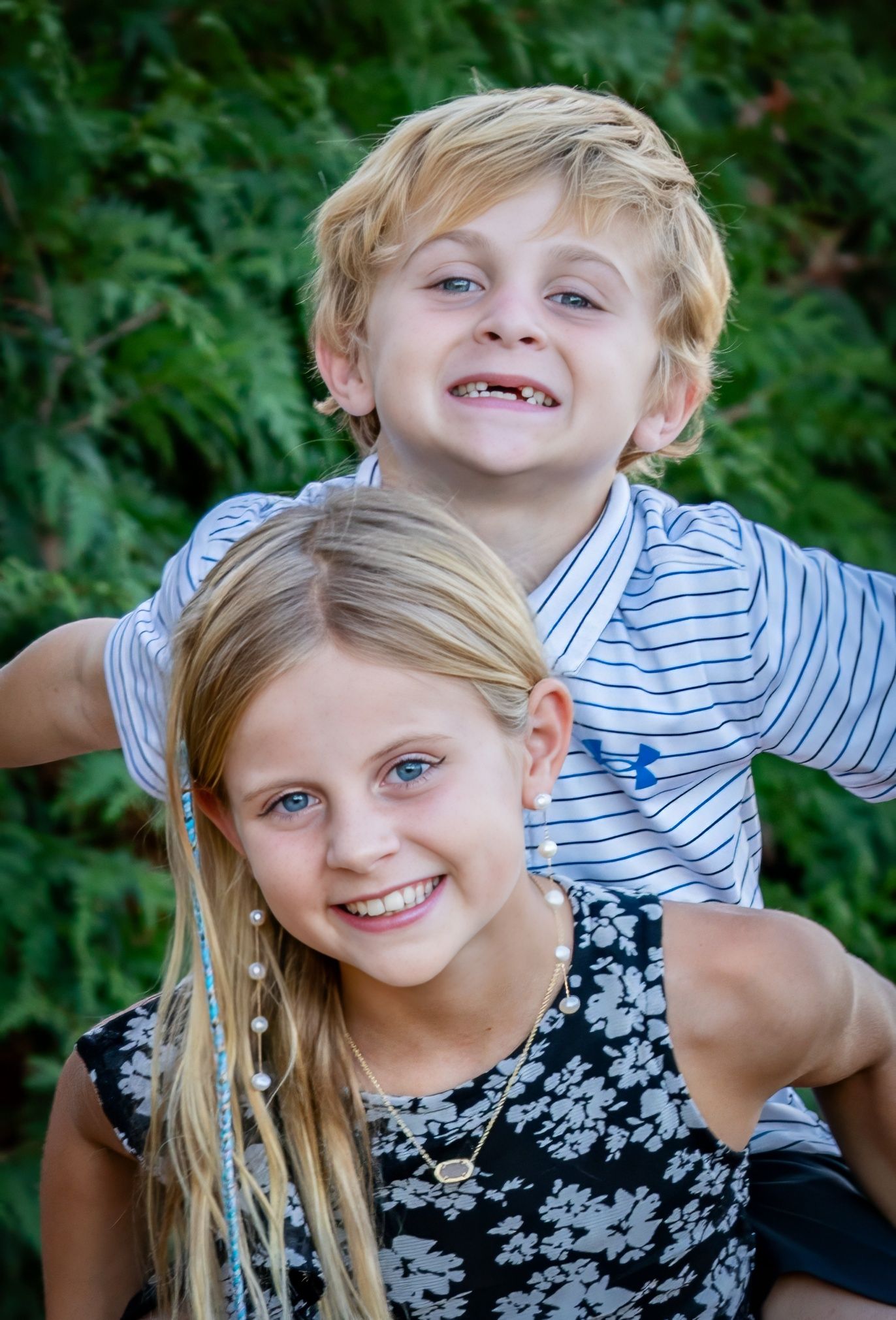 Two blonde siblings smiling outdoors, girl giving boy a piggyback ride in front of green foliage.