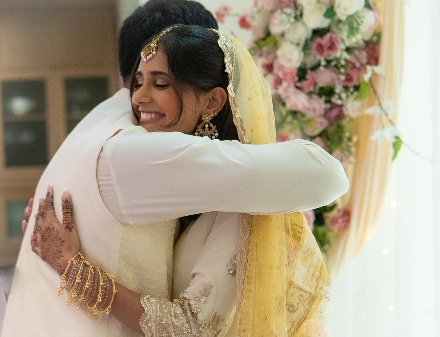 South Asian bride in white wedding attire with henna hands shares warm embrace with guest near floral backdrop.