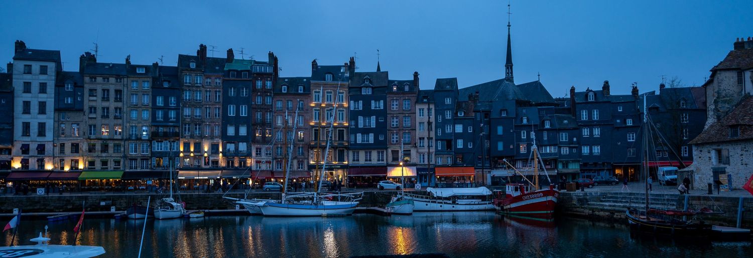 Panoramic view of Honfleur harbor at dusk with illuminated buildings and boats reflecting on calm water.