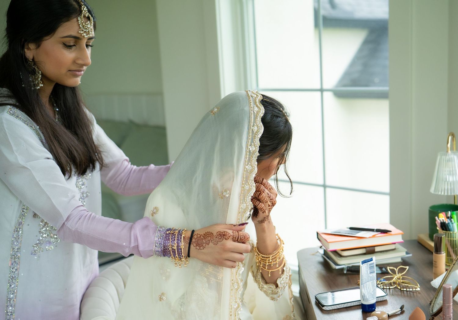 Woman helping South Asian bride adjust white bridal dupatta veil during wedding preparation indoors.