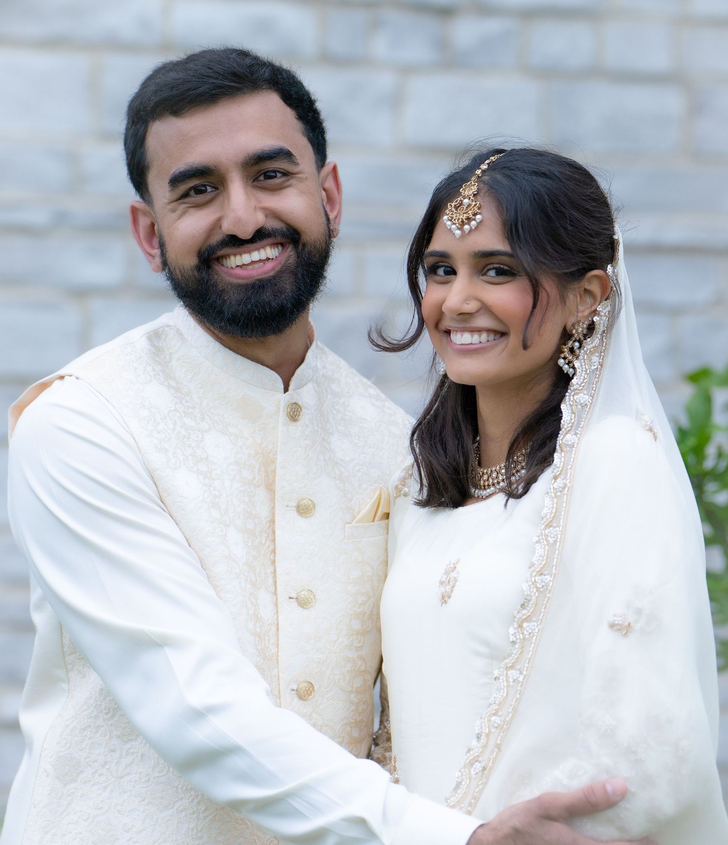 Happy South Asian bride and groom posing together in matching white embroidered wedding outfits.