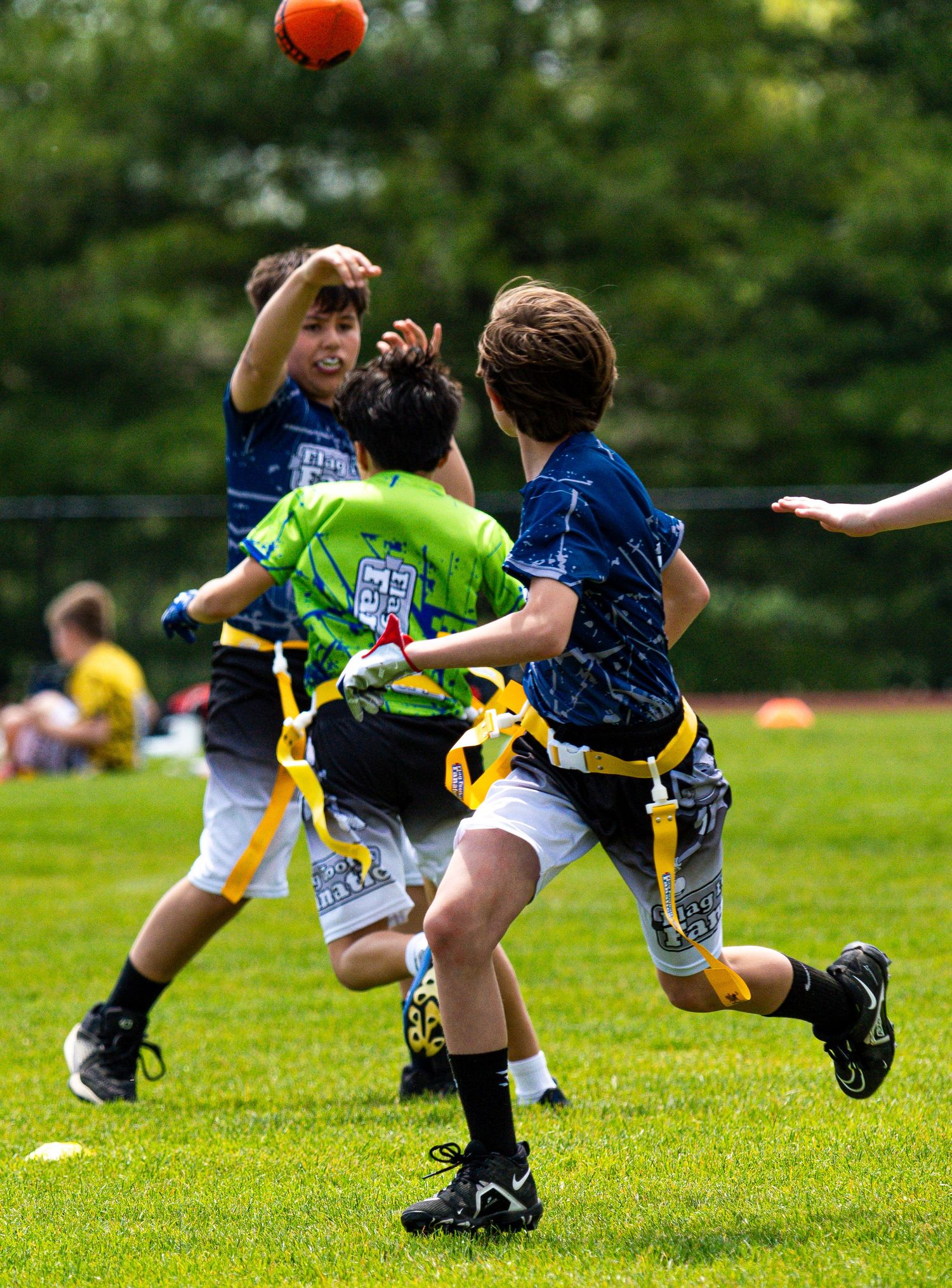 Youth flag football players in blue and green jerseys compete for orange ball on grass field.