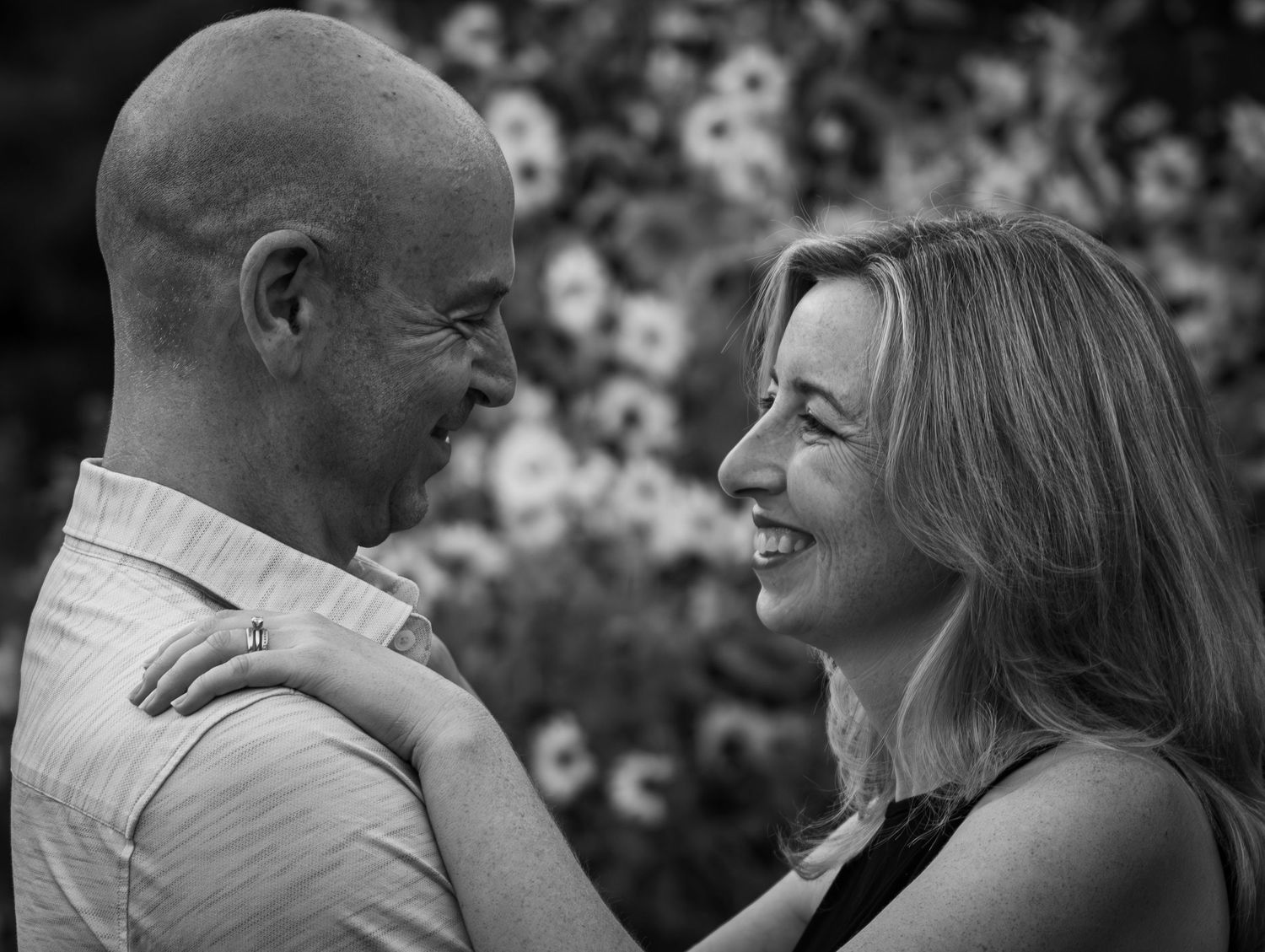 Smiling couple gazing at each other in black and white portrait with flowers behind.