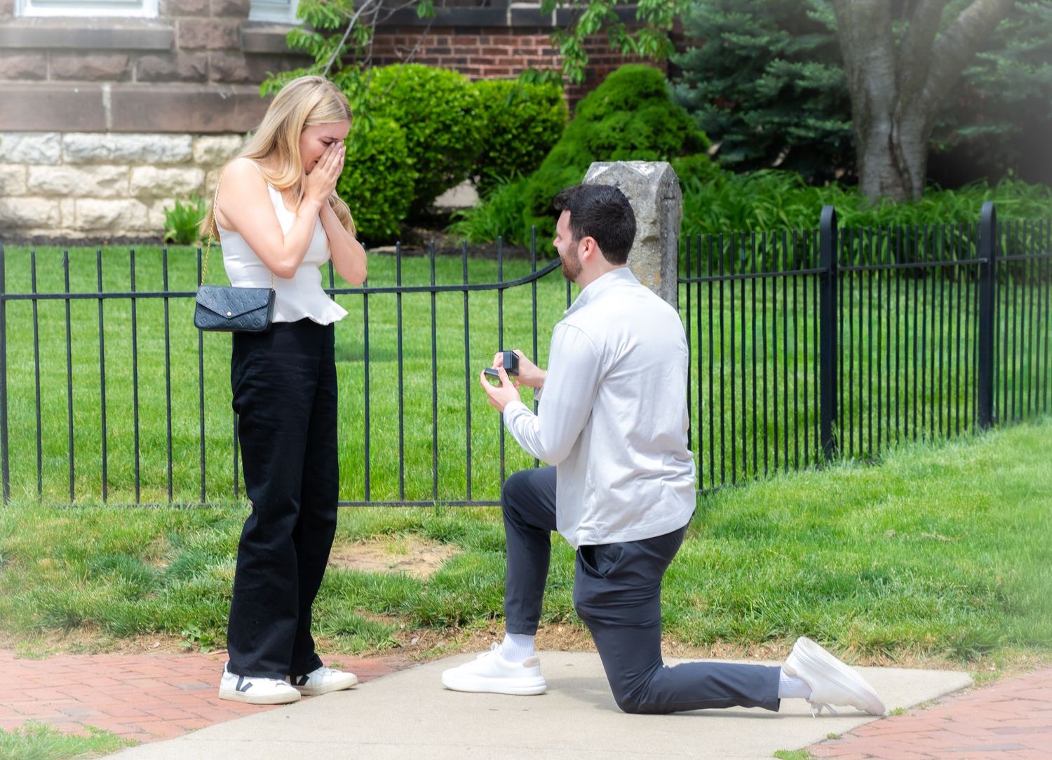 Man proposes on one knee to emotional blonde woman outdoors near iron fence on sunny day.