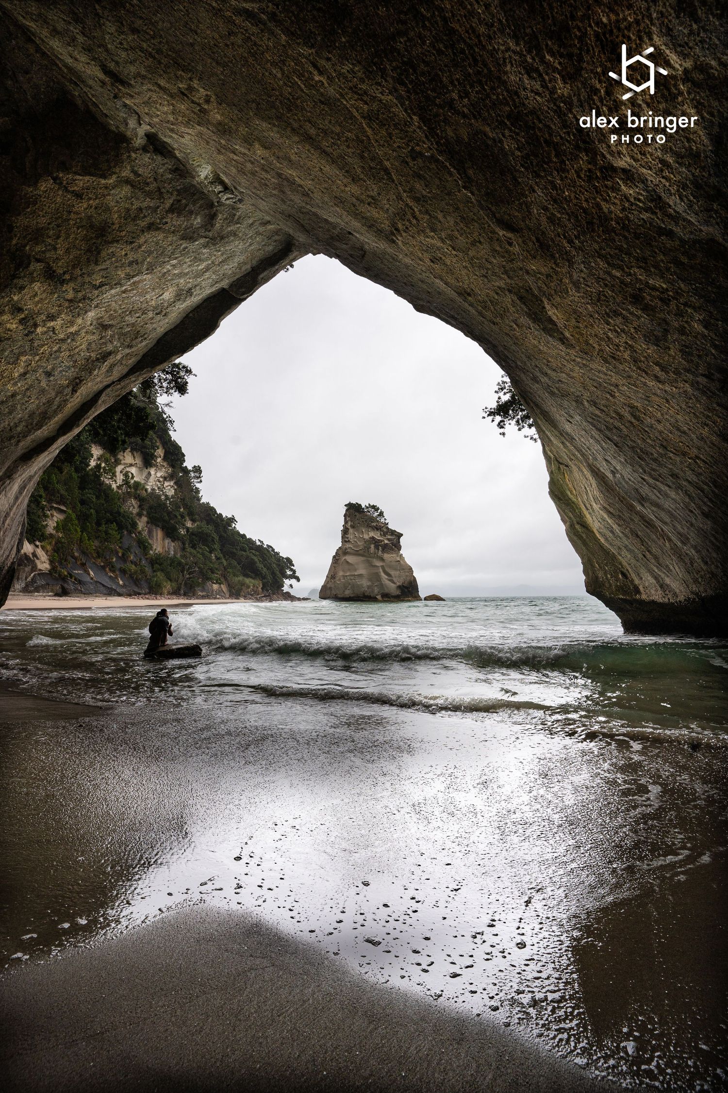 A person sits on a sandy beach viewed through a dramatic natural rock arch with a sea stack in the ocean.