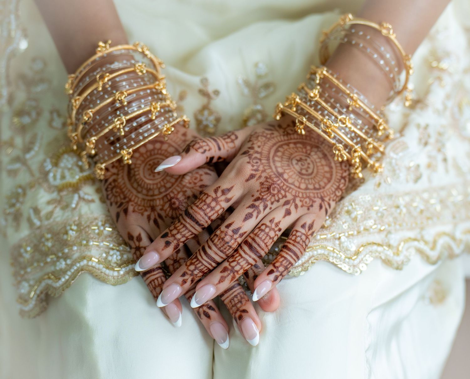 Bride's hands adorned with intricate henna mehndi designs and gold bangles on white wedding dress.