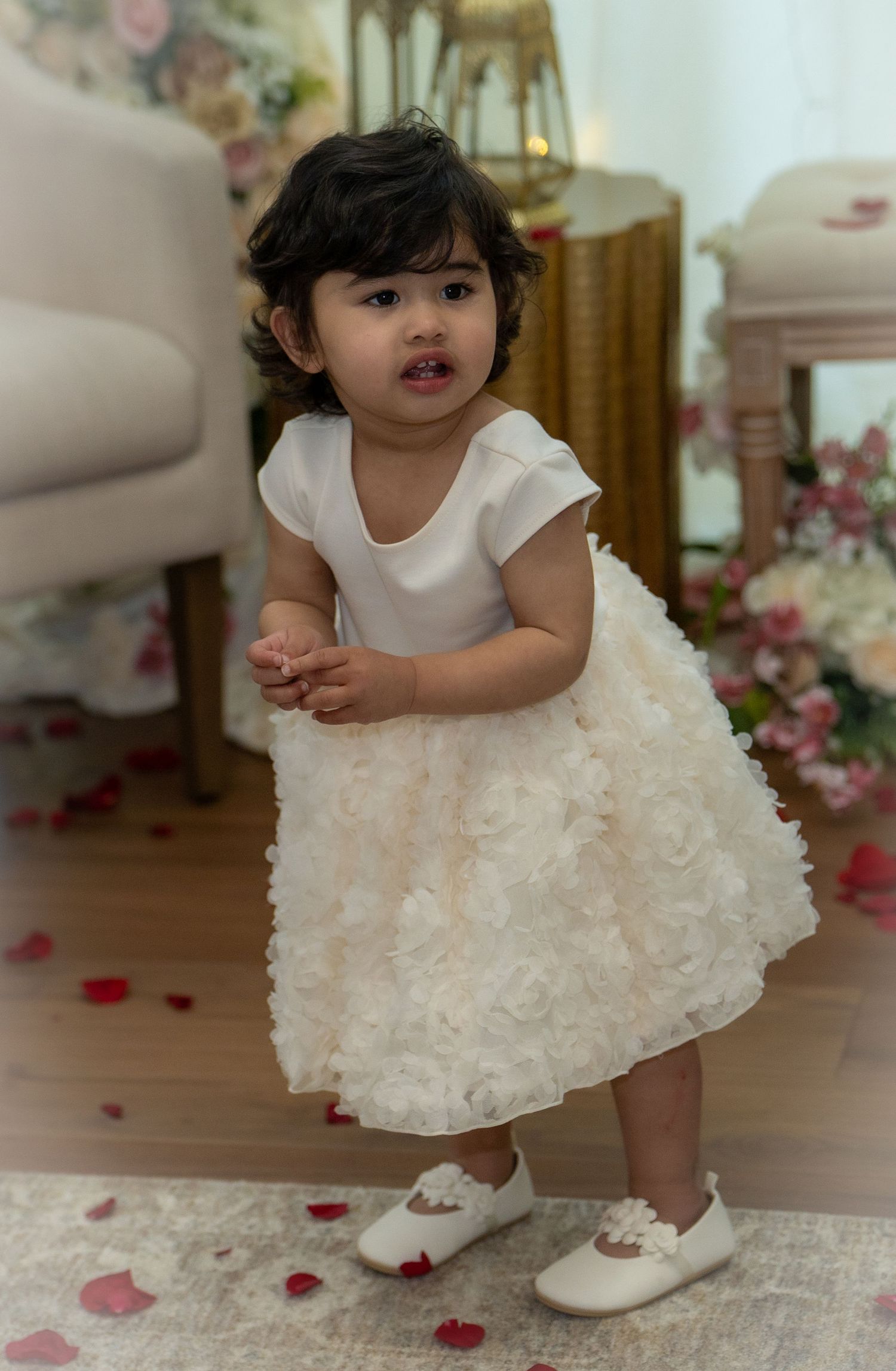 Cute toddler in white floral dress standing among rose petals indoors.