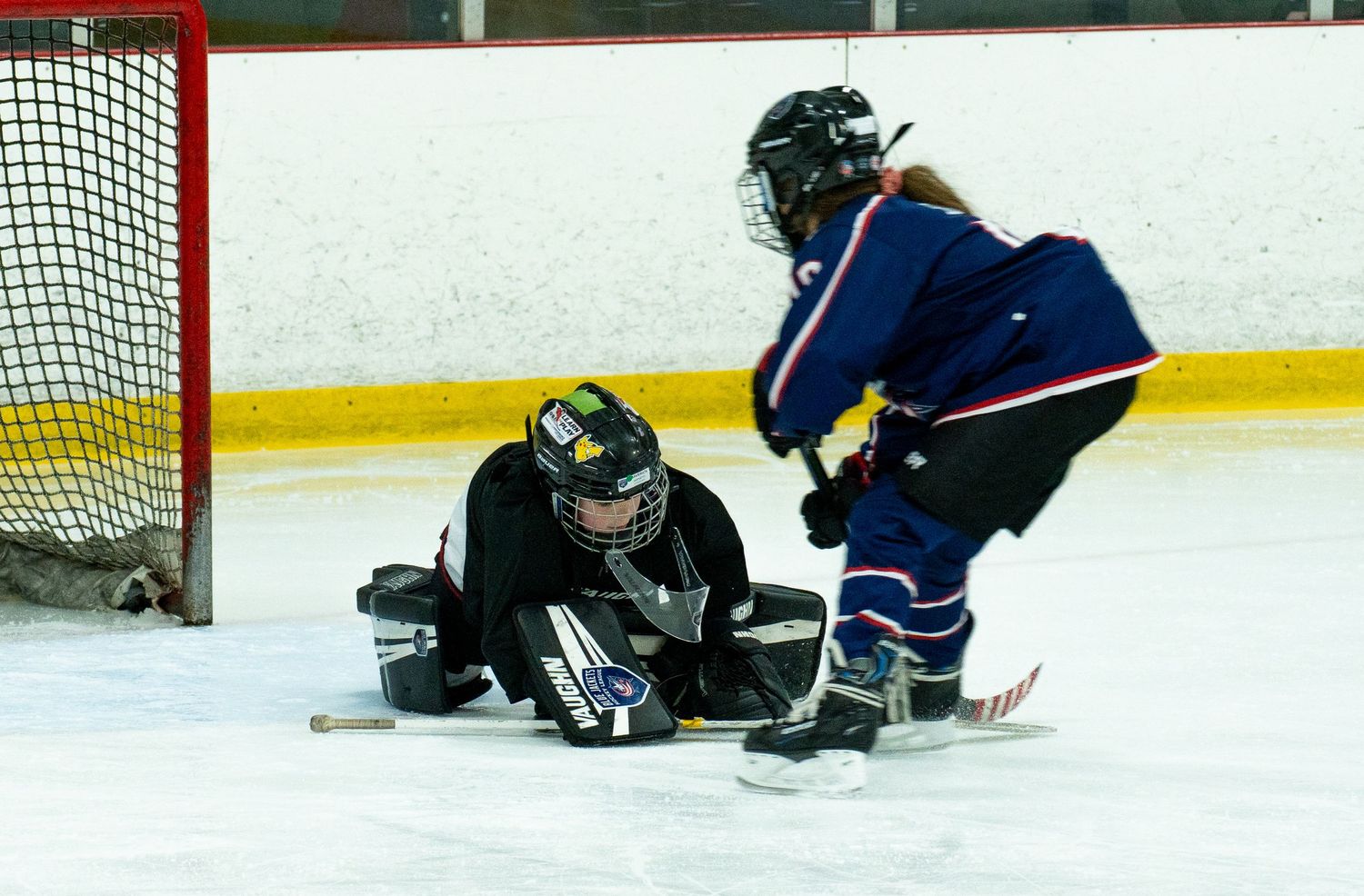 Youth hockey goalie making save against opposing player during indoor ice hockey game.
