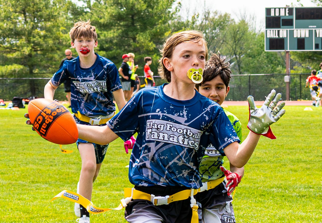 Kids playing flag football outdoors, with one boy carrying an orange football while wearing blue team jerseys.