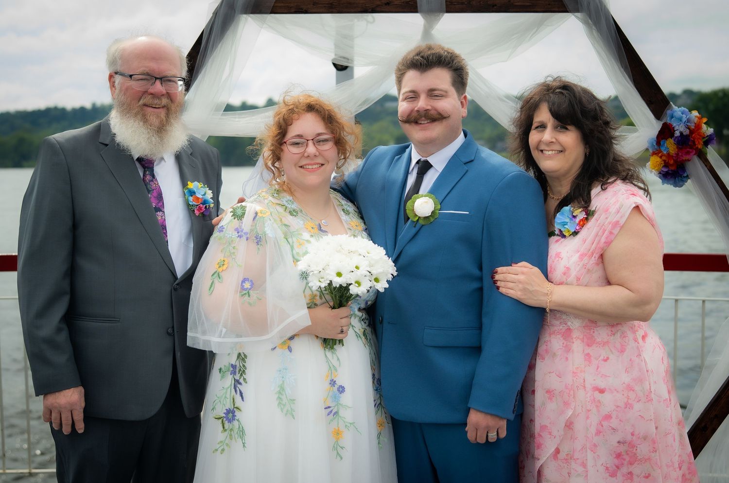 Two couples pose together at an outdoor waterfront wedding ceremony, bride holding white flowers.