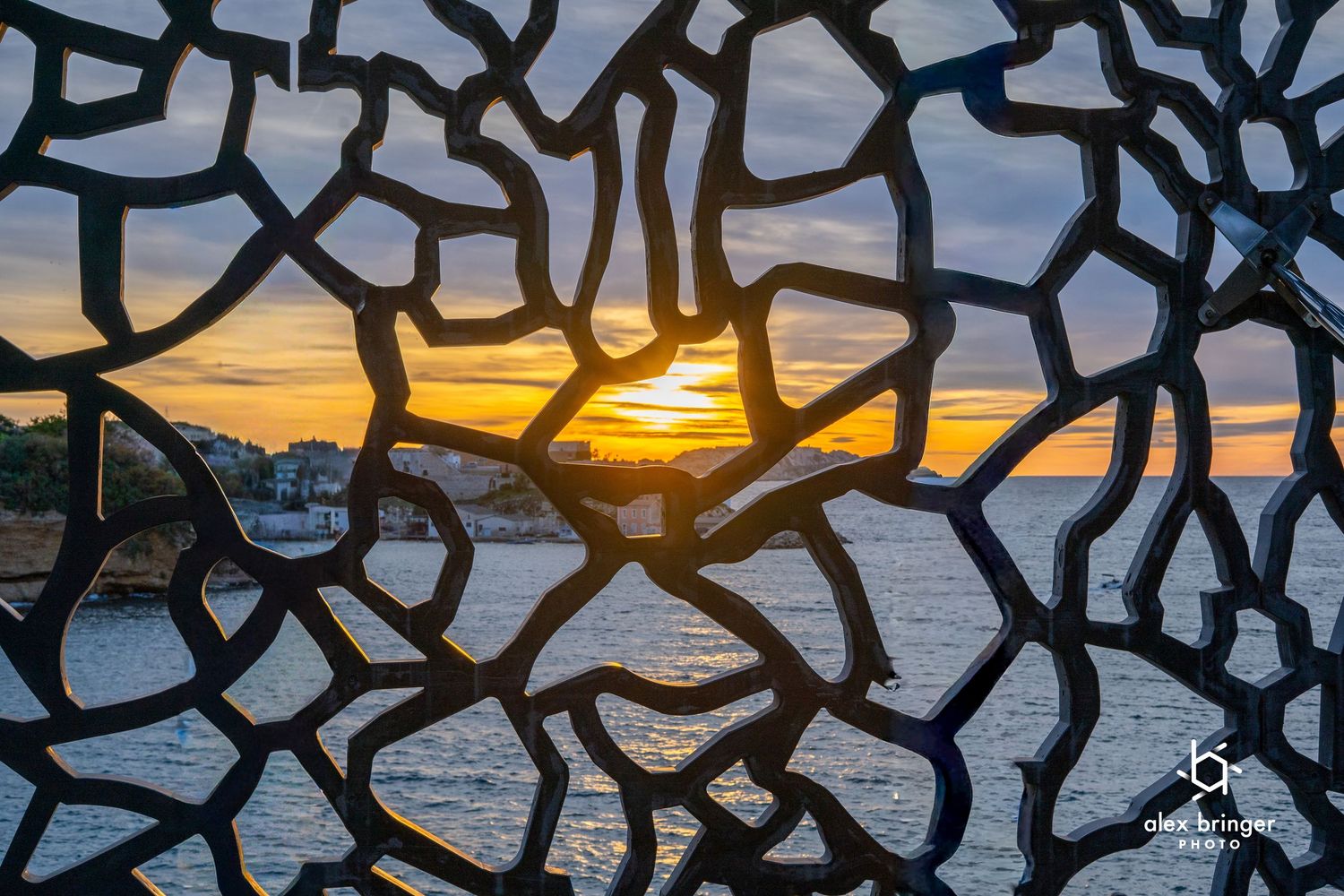 Sunset viewed through intricate metal lattice sculpture overlooking a calm ocean coastline.