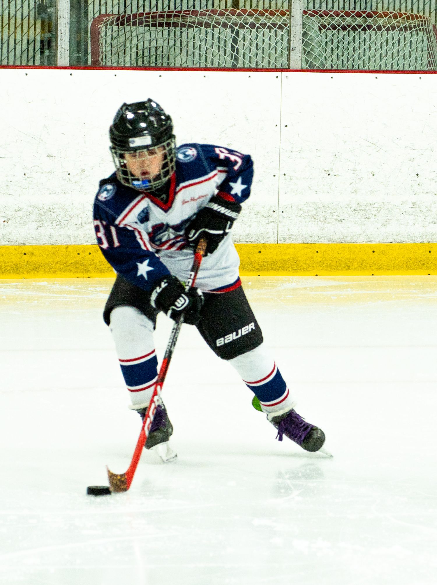 Youth hockey player in blue and white Bauer uniform controls puck on ice rink during game.