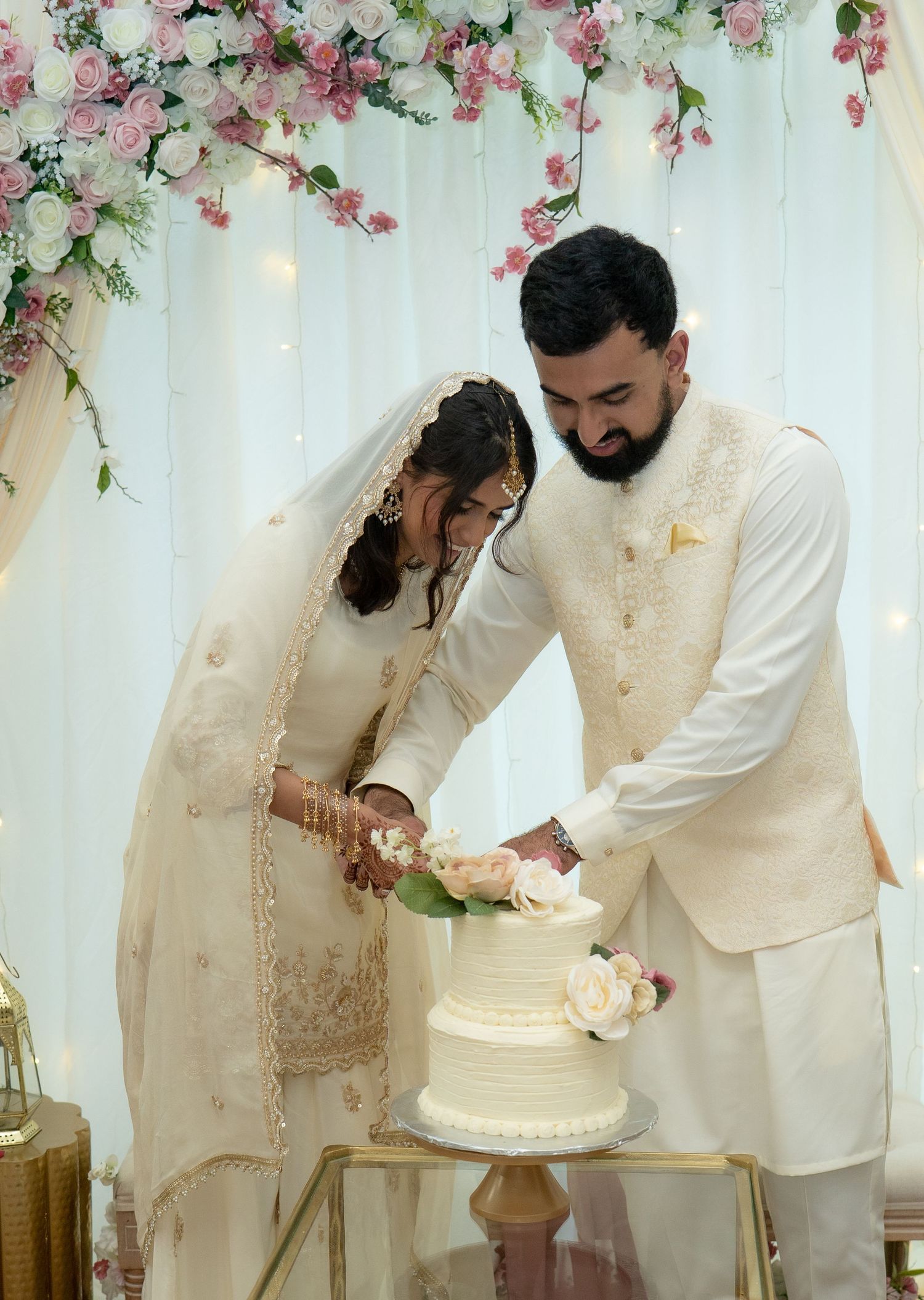 South Asian bride and groom in white traditional attire cut wedding cake under pink floral arch.