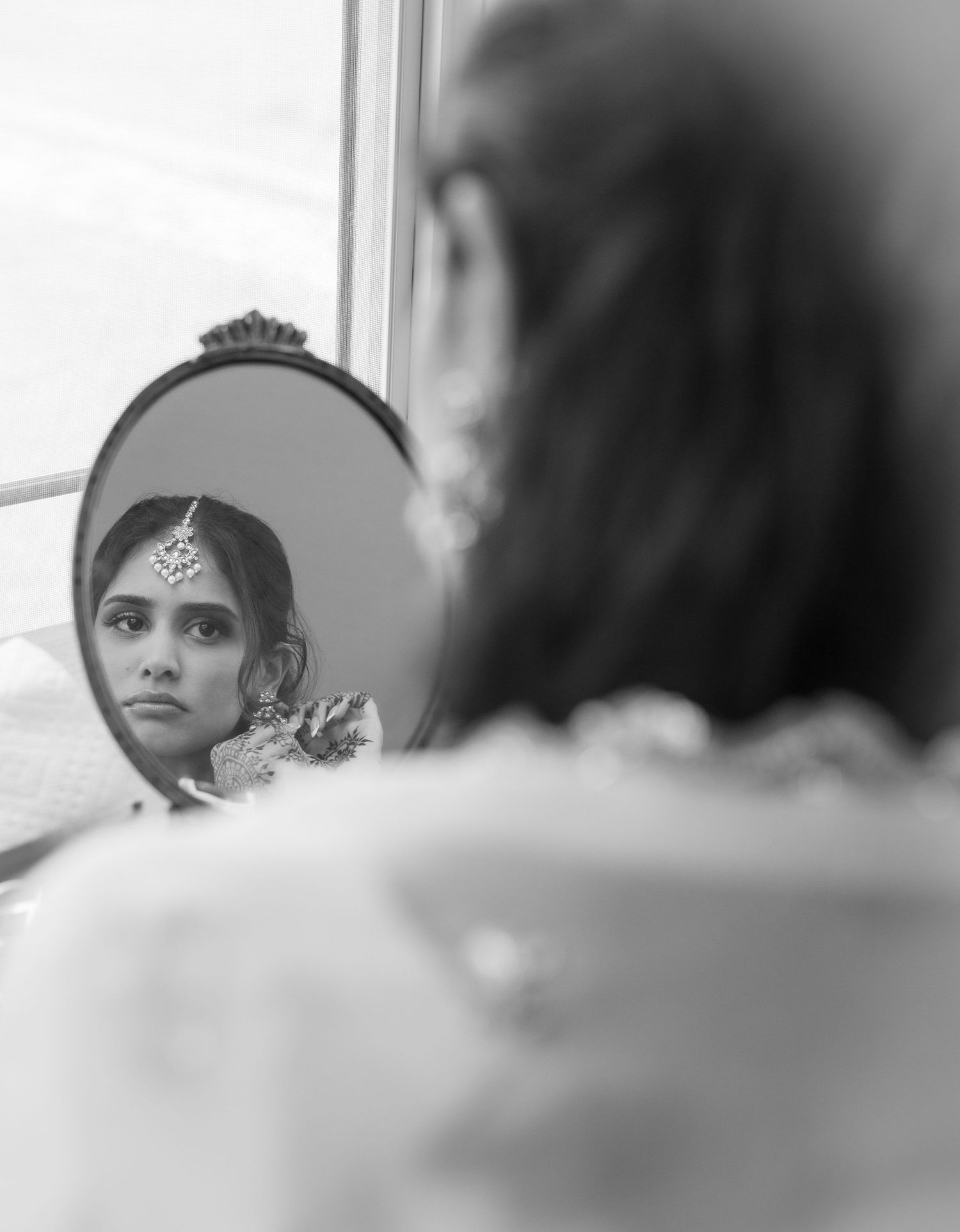 Indian bride gazes at her reflection in an ornate oval mirror wearing traditional bridal jewelry in black and white photo.