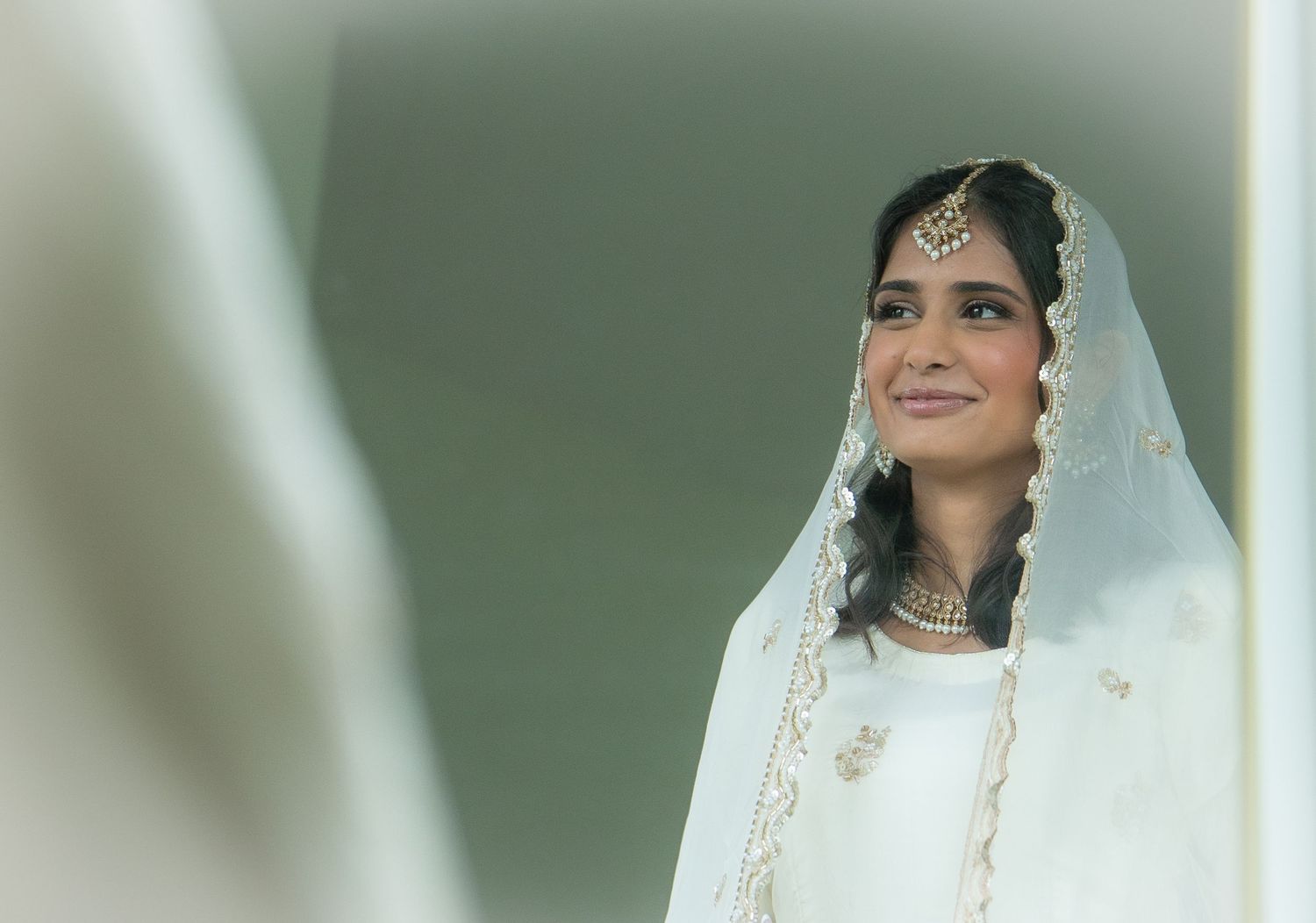 South Asian bride in white wedding dupatta and traditional jewelry smiling softly in mirror reflection.