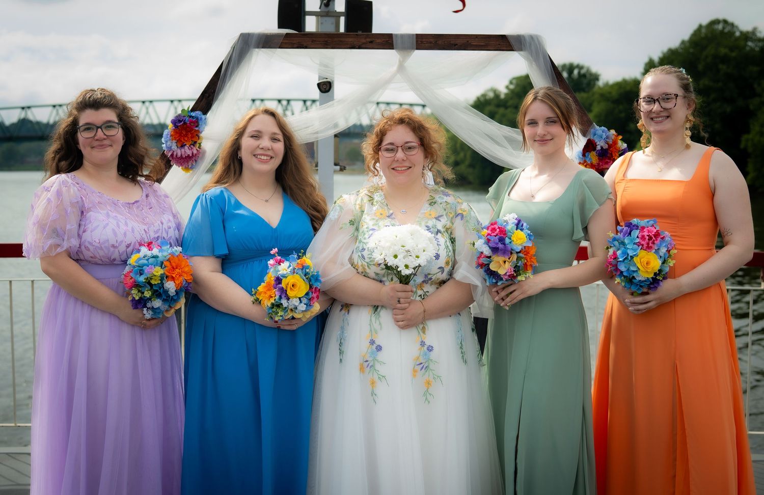 Bride in white gown with four bridesmaids in colorful dresses holding bouquets at an outdoor waterfront wedding.