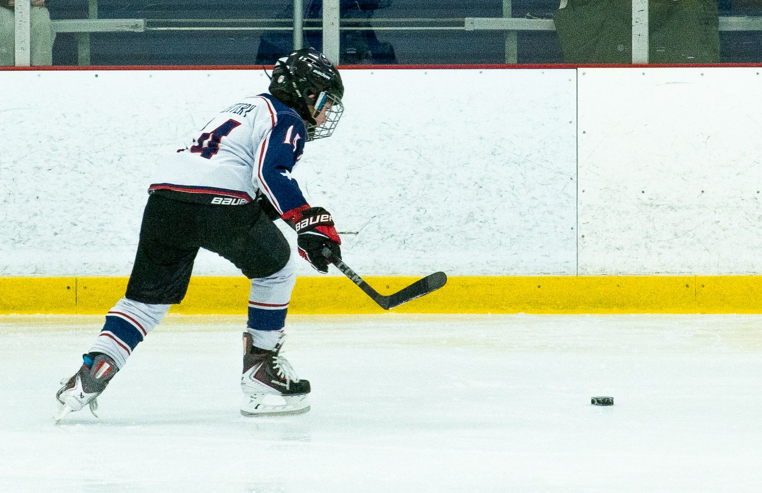 Youth hockey player in navy uniform skating and shooting puck on indoor ice rink.
