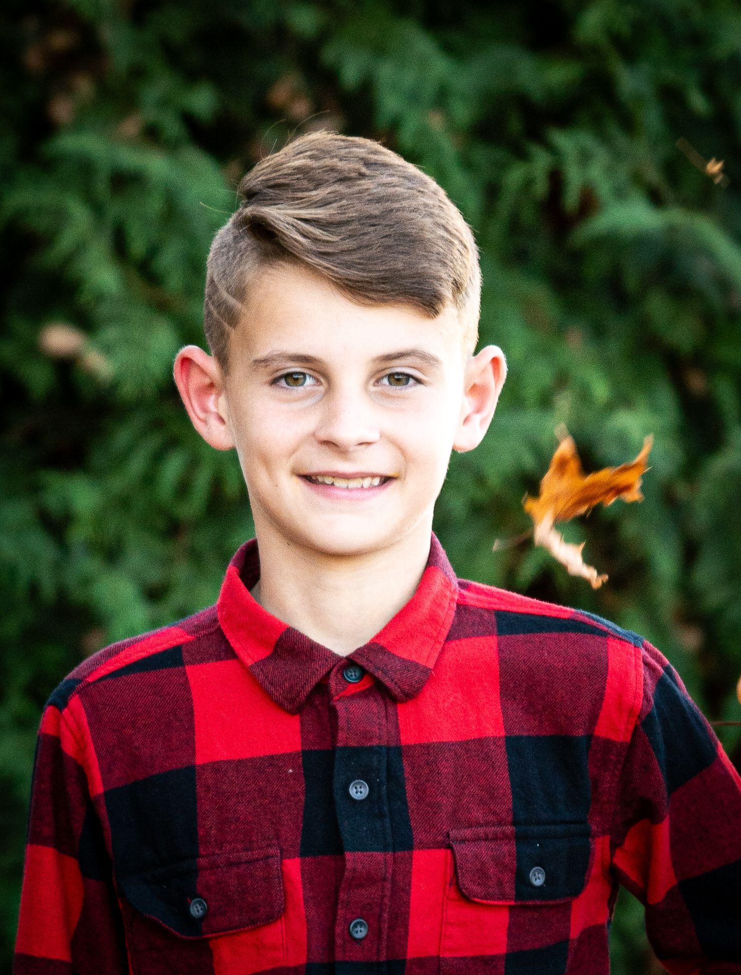 Smiling preteen boy in red plaid flannel shirt with autumn leaf flying in background outdoors.