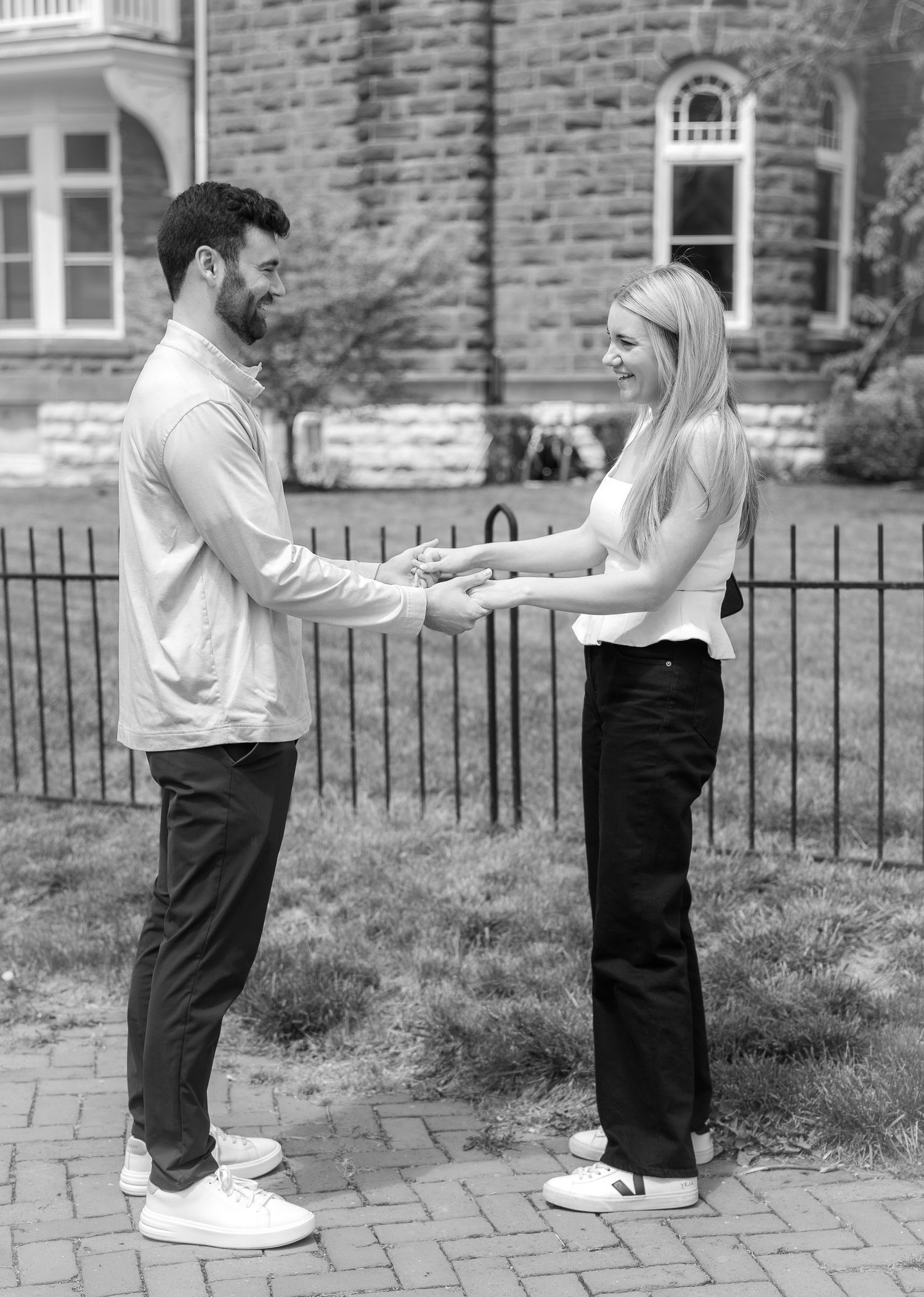 Black and white photo of engaged couple holding hands and smiling on brick pathway.