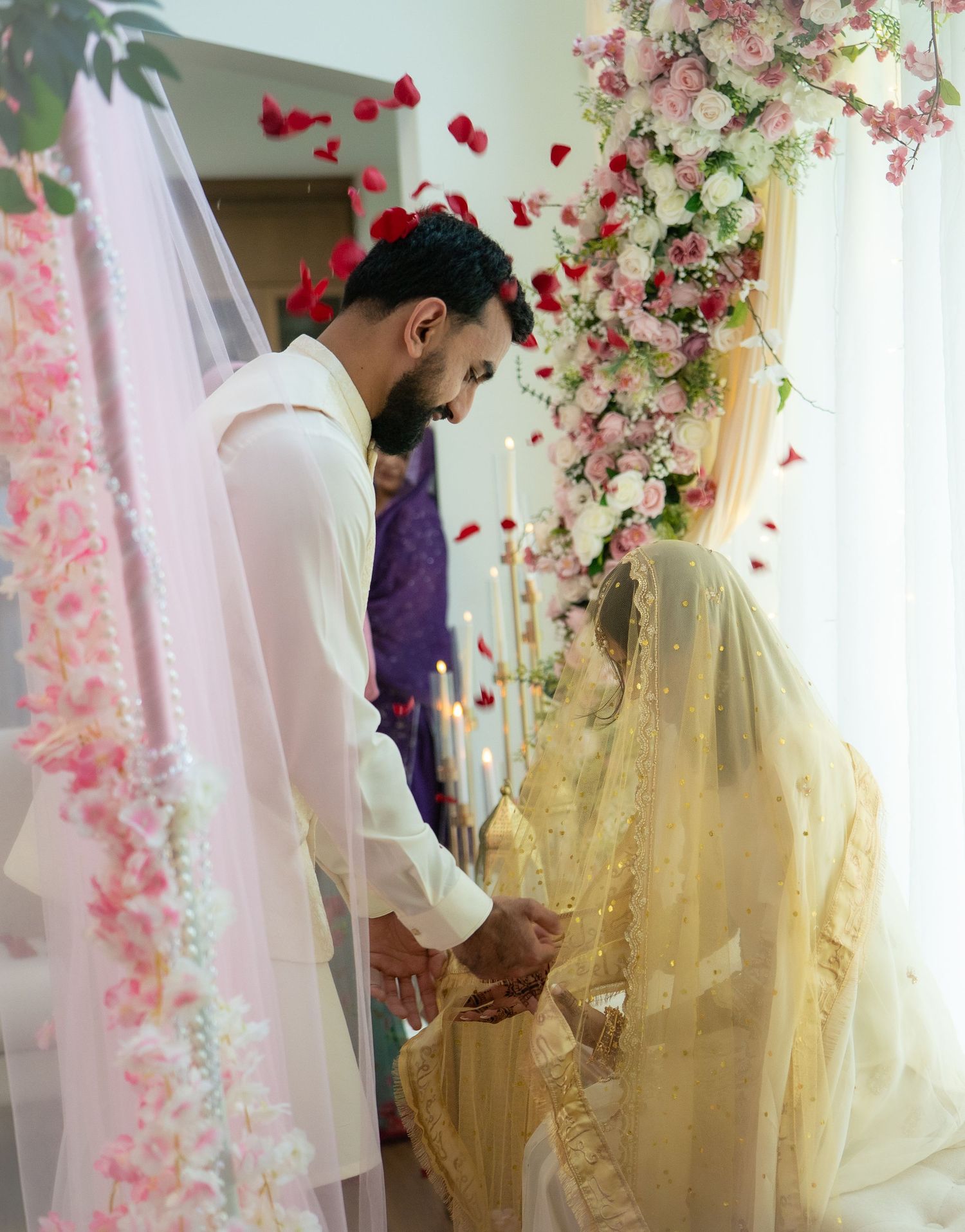 Muslim couple holding hands during Nikah ceremony with pink floral backdrop and rose petals falling.