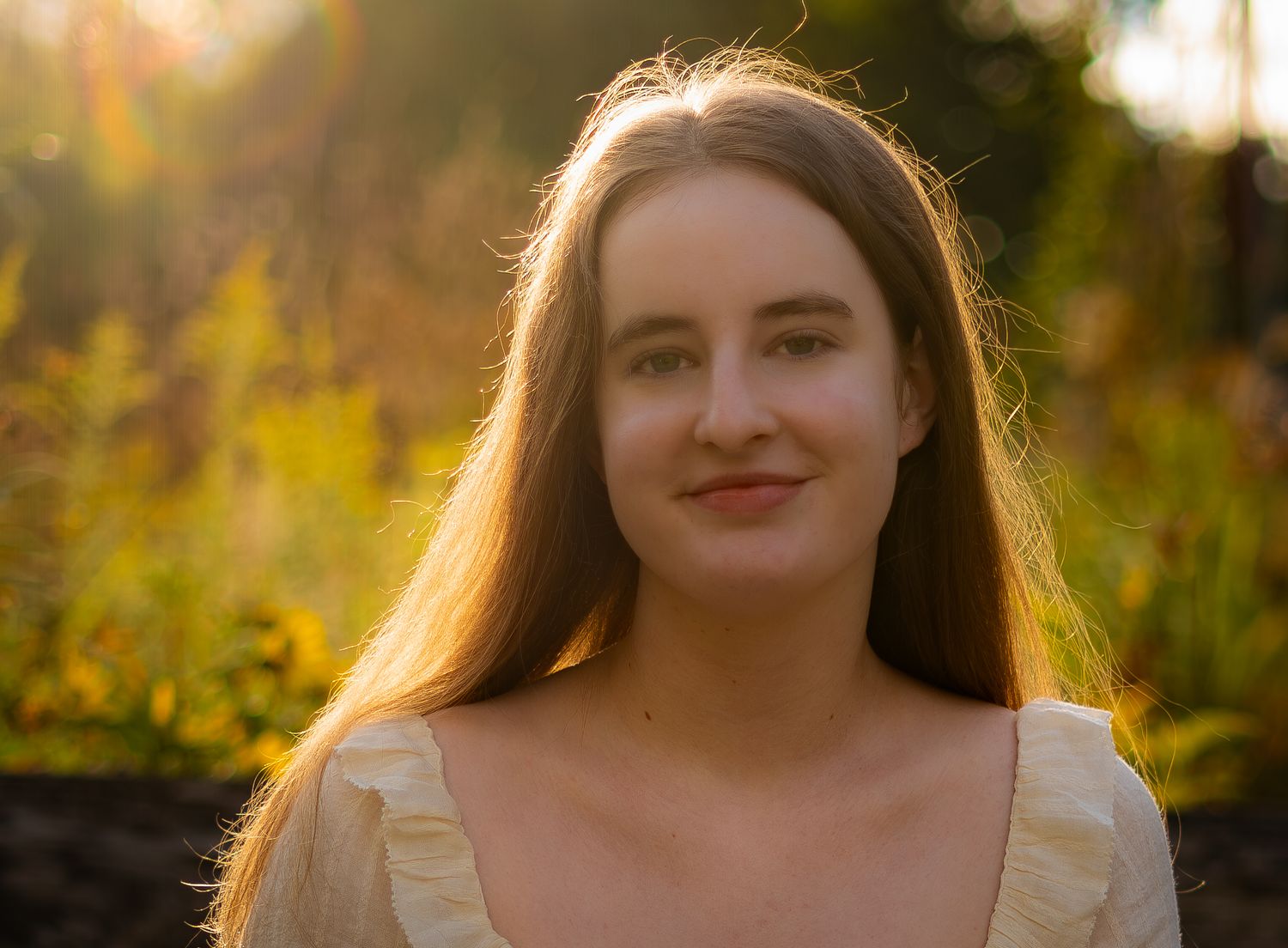 Young woman with long brown hair smiling in golden sunset light outdoors.