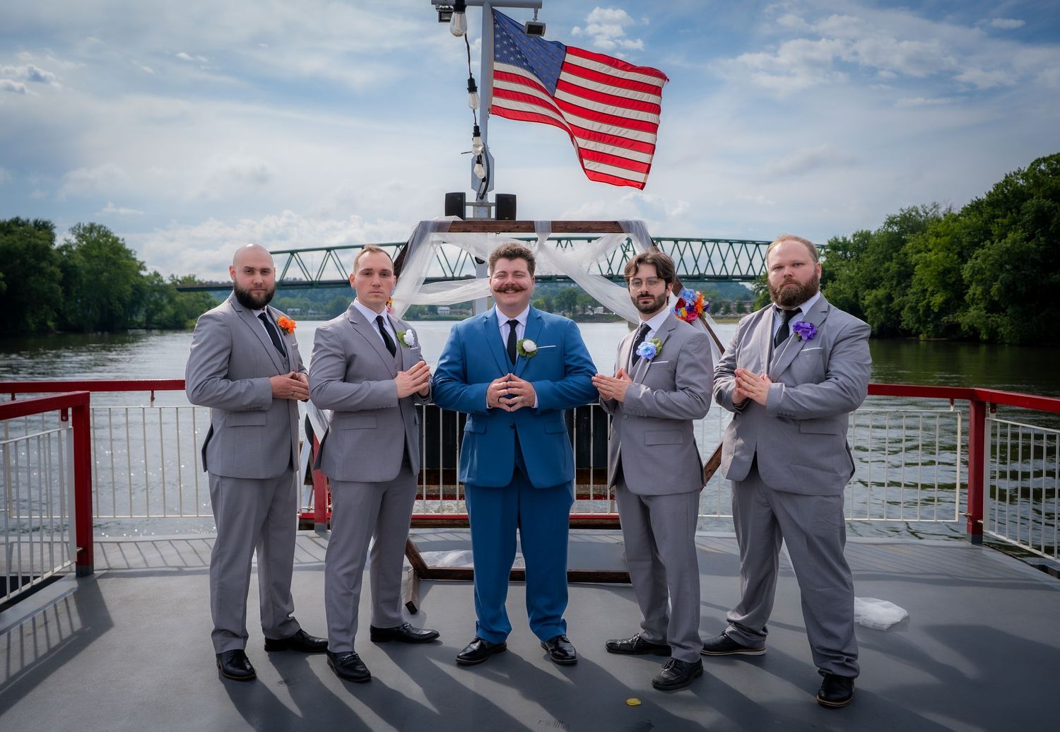 Groom in blue suit with groomsmen in gray suits posing on a riverboat deck with American flag backdrop.