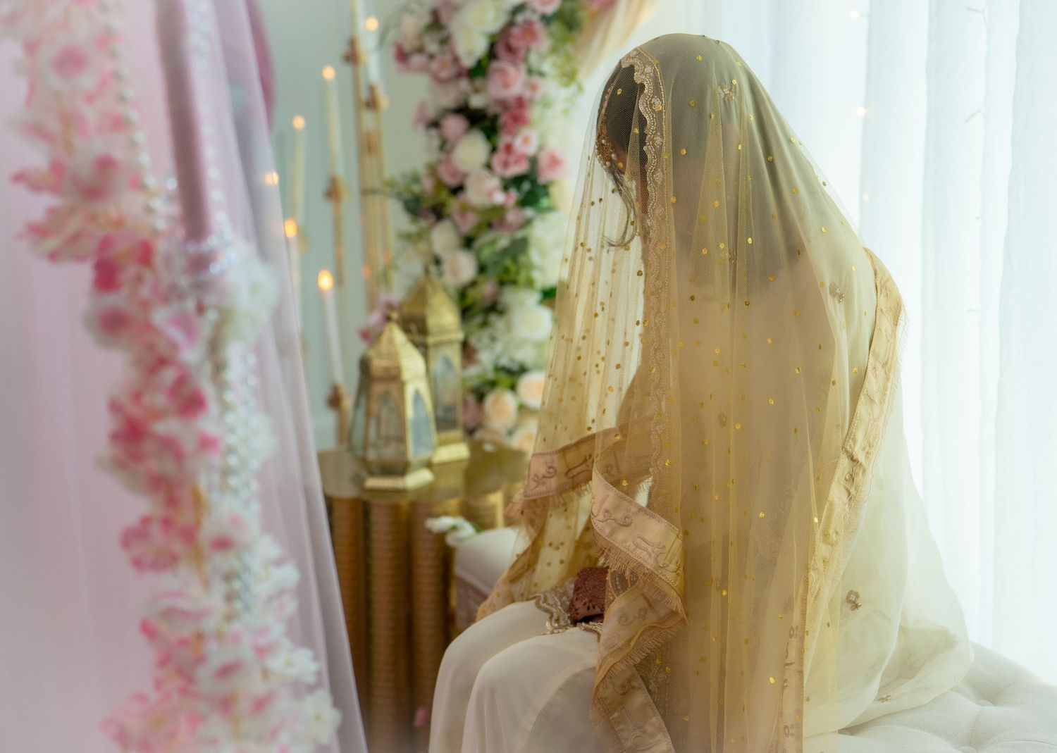 Muslim bride in gold embroidered veil seated before floral arrangement during Nikah ceremony.