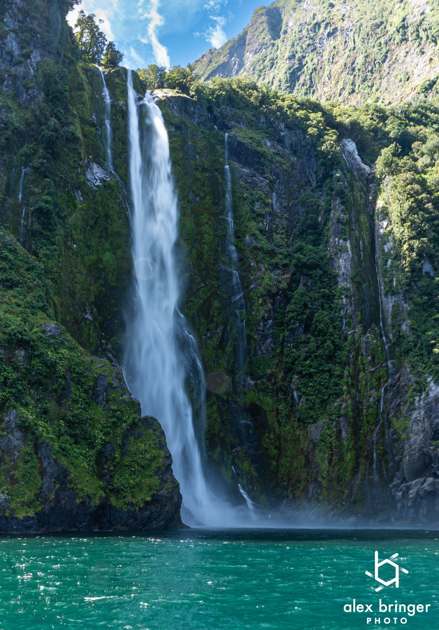 Massive waterfall cascades down lush green cliffs into turquoise waters at Milford Sound, New Zealand.