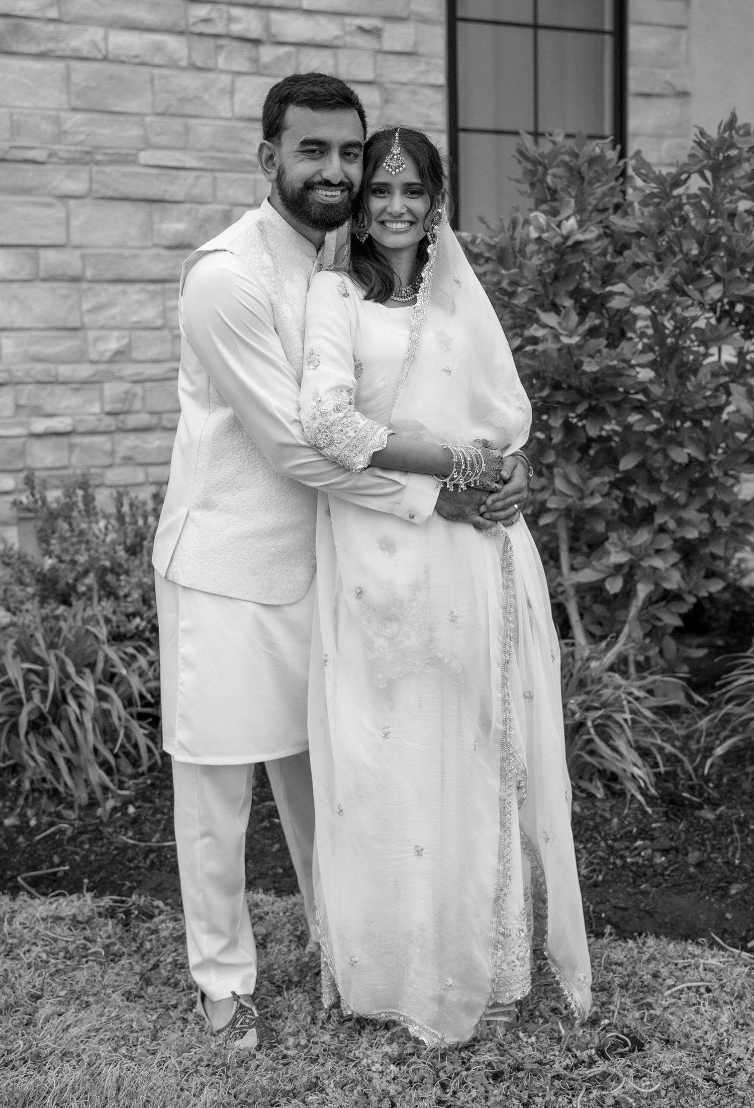 Indian couple in traditional white wedding attire embracing outdoors in a black and white portrait photo.