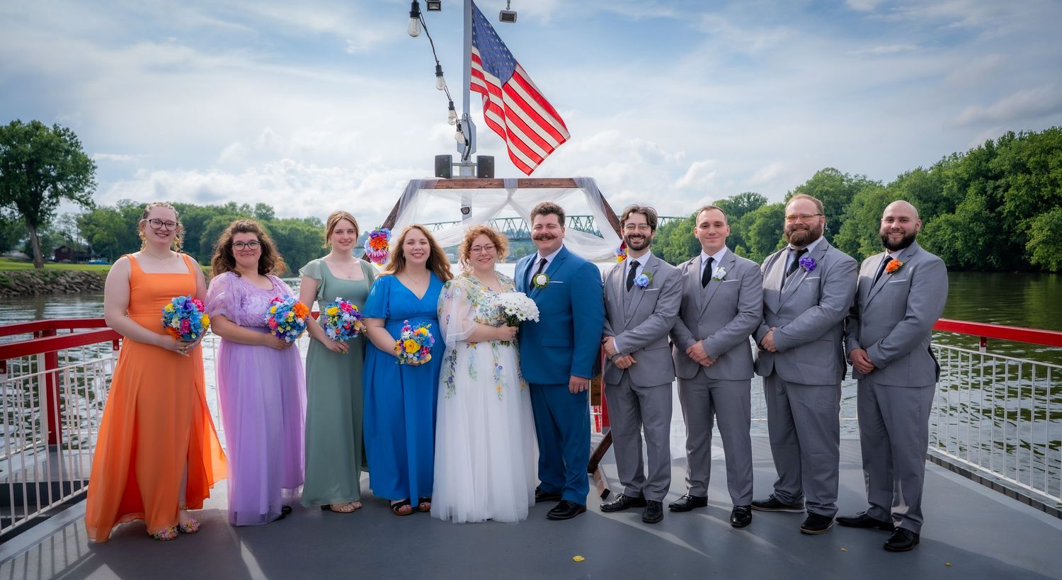 Wedding party poses on a boat deck with an American flag, bridesmaids in colorful gowns and groomsmen in gray suits.