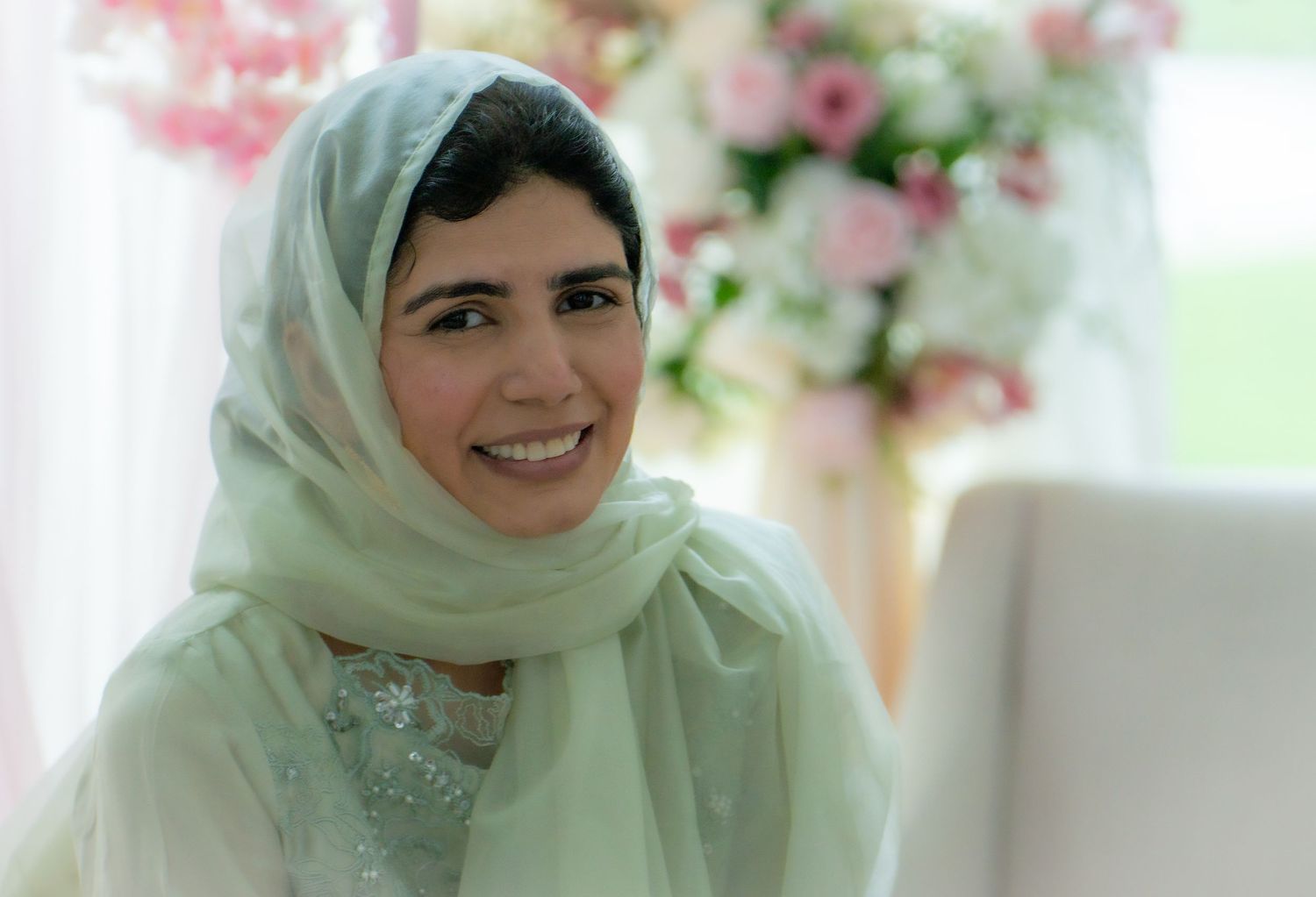 Smiling woman in white hijab seated near pink floral arrangement.