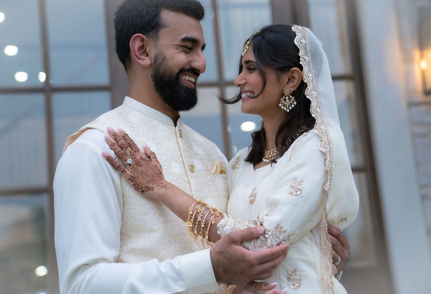An Indian couple in traditional white and gold wedding attire smiling warmly at each other.
