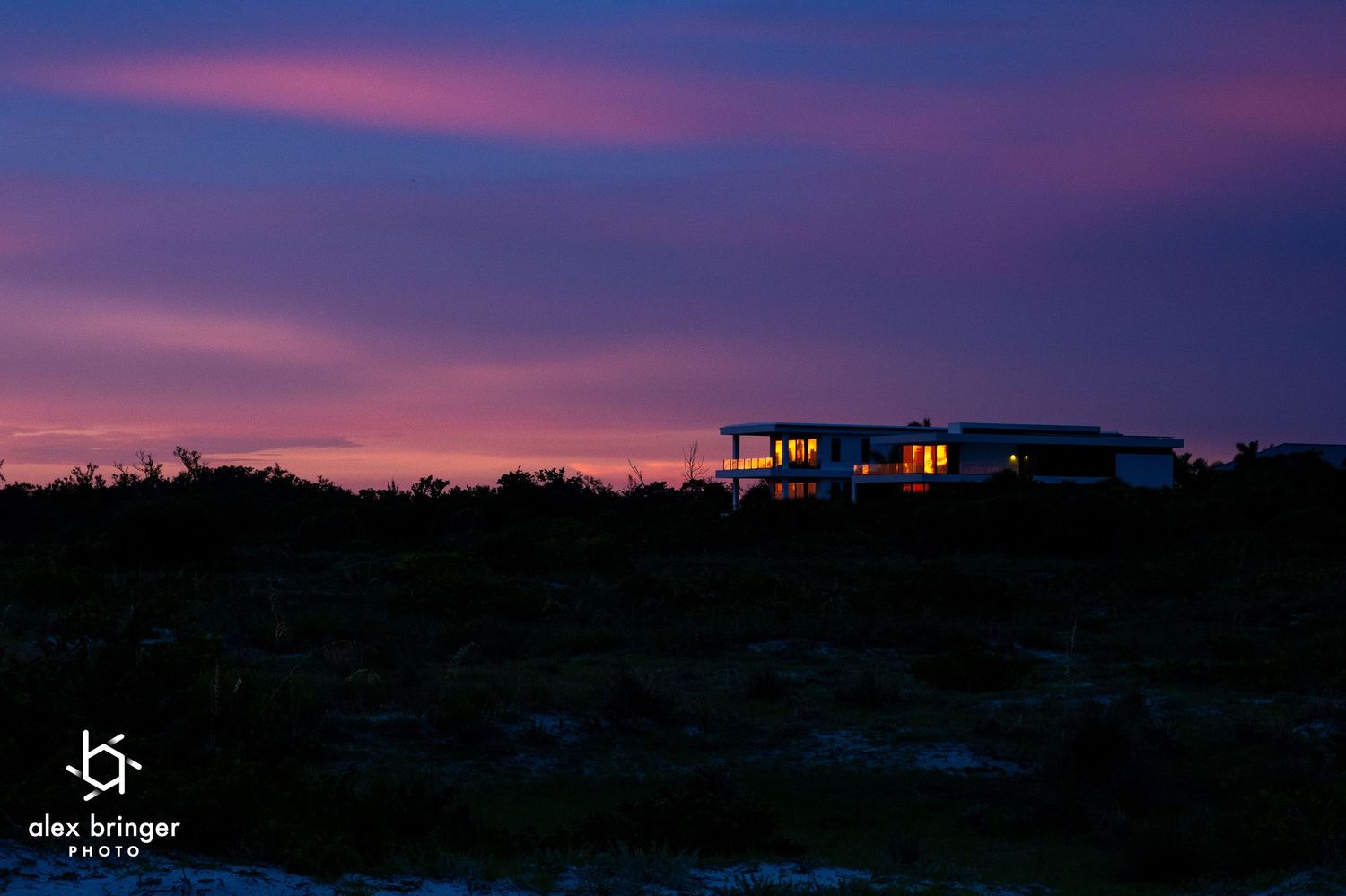 A modern house illuminated against a stunning purple and pink sunset sky over open wilderness.