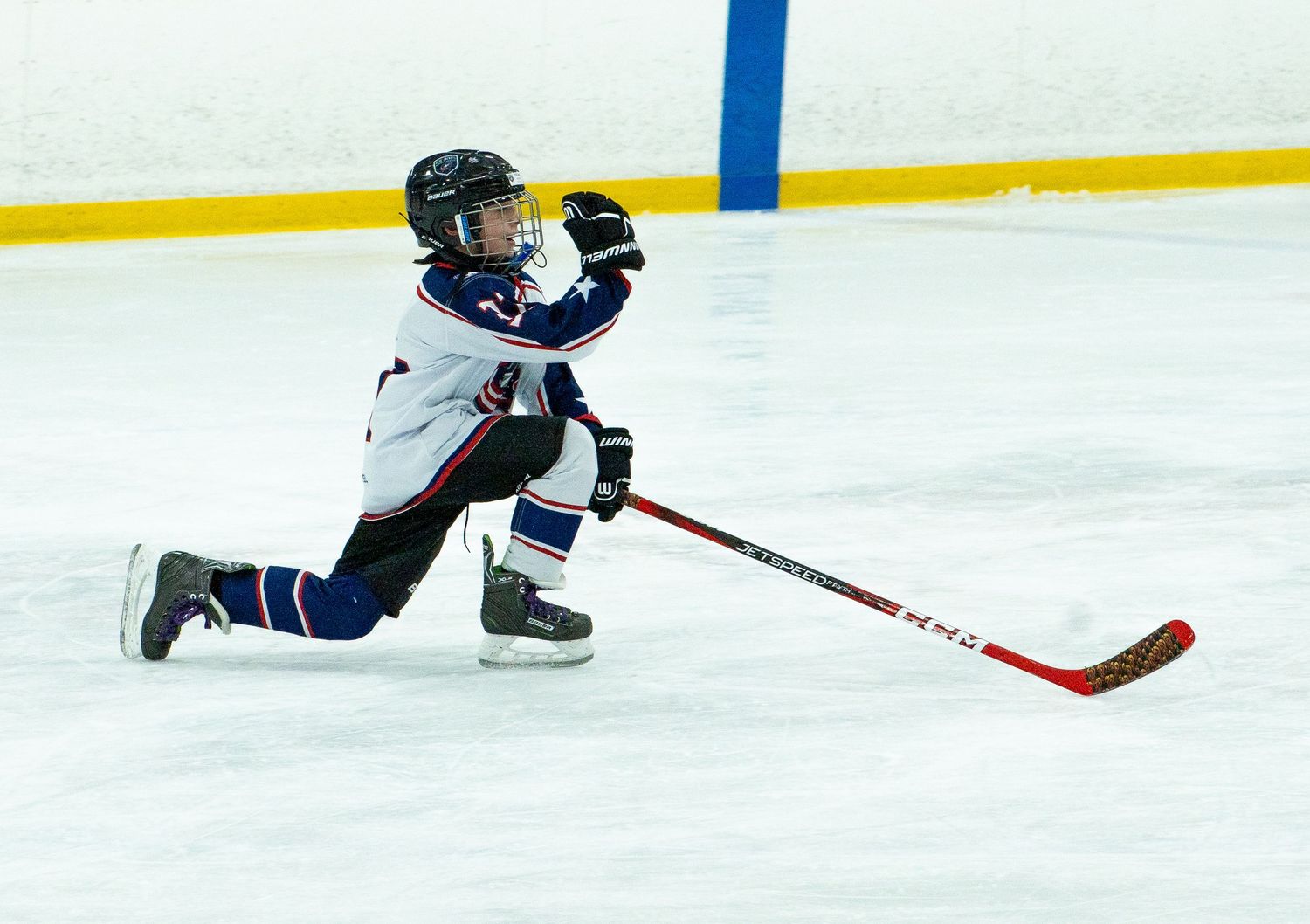 Young hockey player slides on ice in white and blue uniform during a youth ice hockey game.
