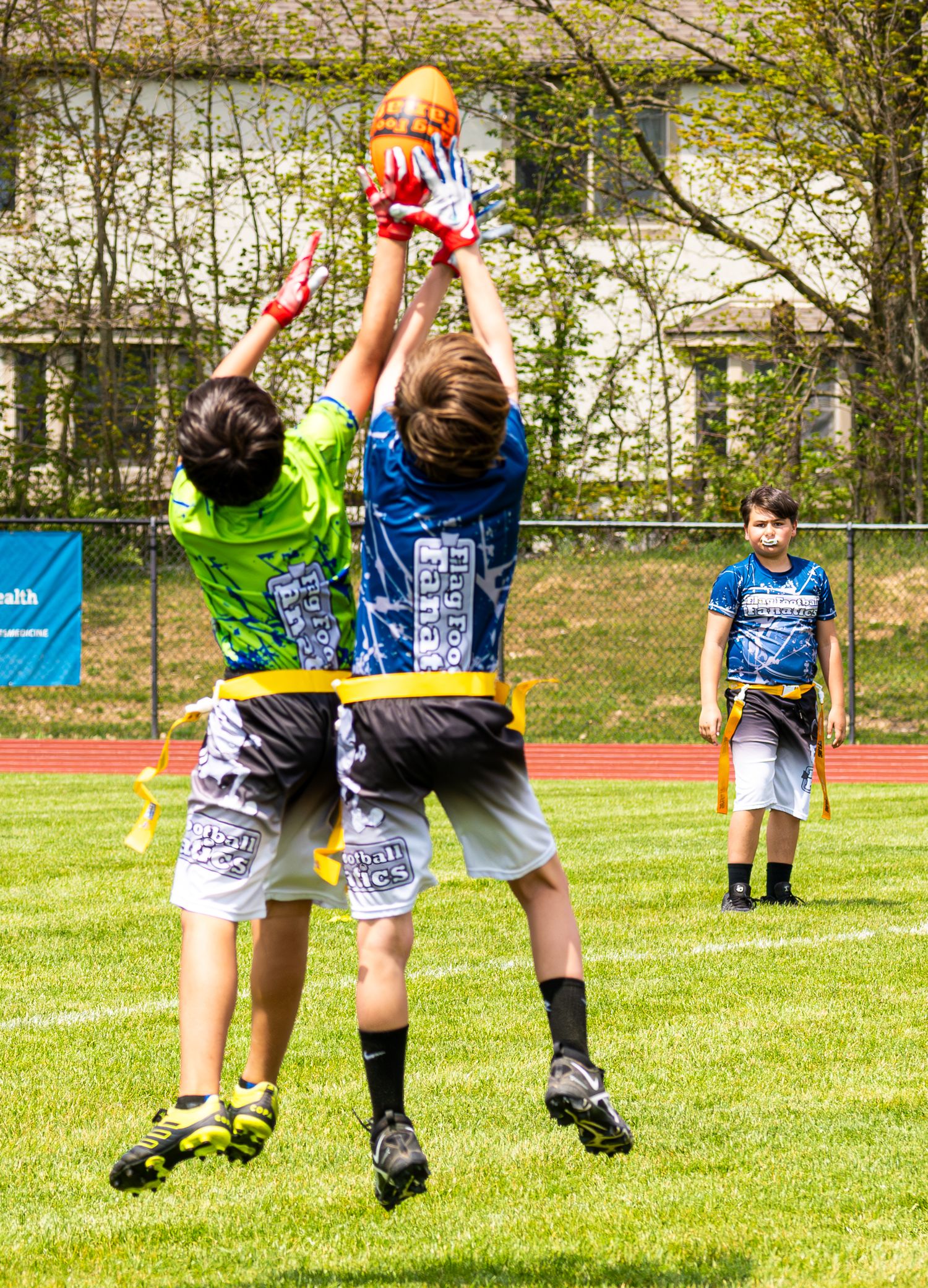 Two youth flag football players jump to catch an orange football during an outdoor game on green field.