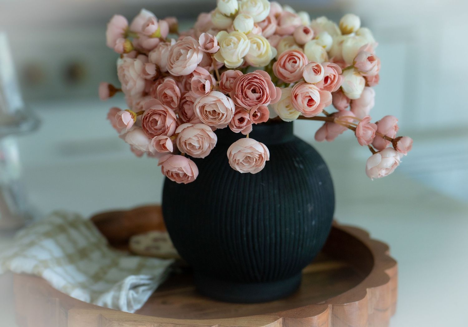 Pink and white roses arranged in a dark ribbed round vase on a wooden tray.