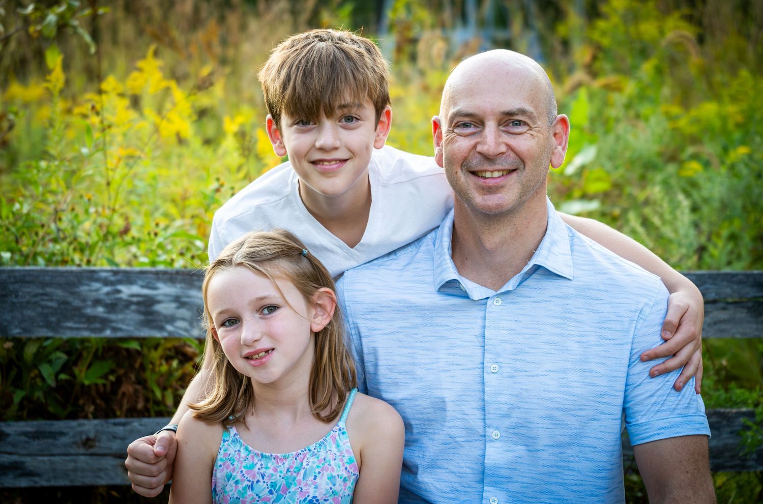 Father sitting on bench smiling with son and daughter in outdoor family portrait.