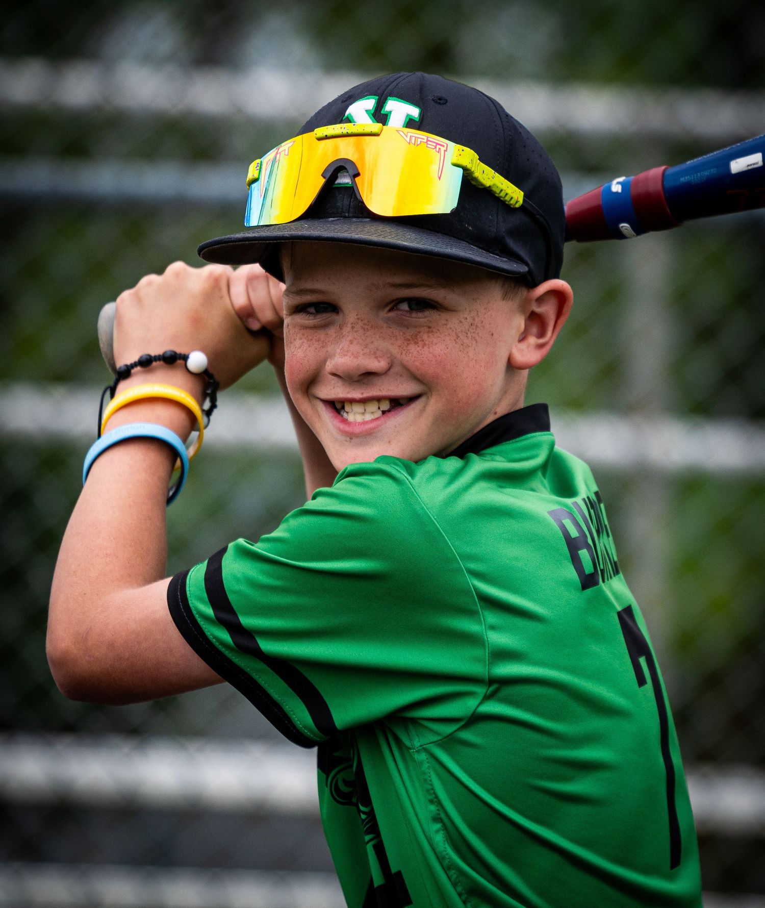 Young baseball player in green uniform smiles wearing yellow sunglasses and black cap at bat.