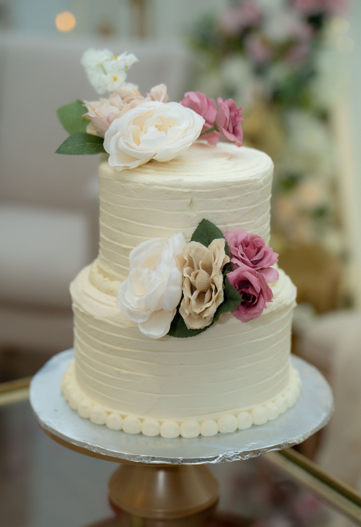 Elegant two-tier white buttercream wedding cake decorated with pink and cream roses on gold stand.